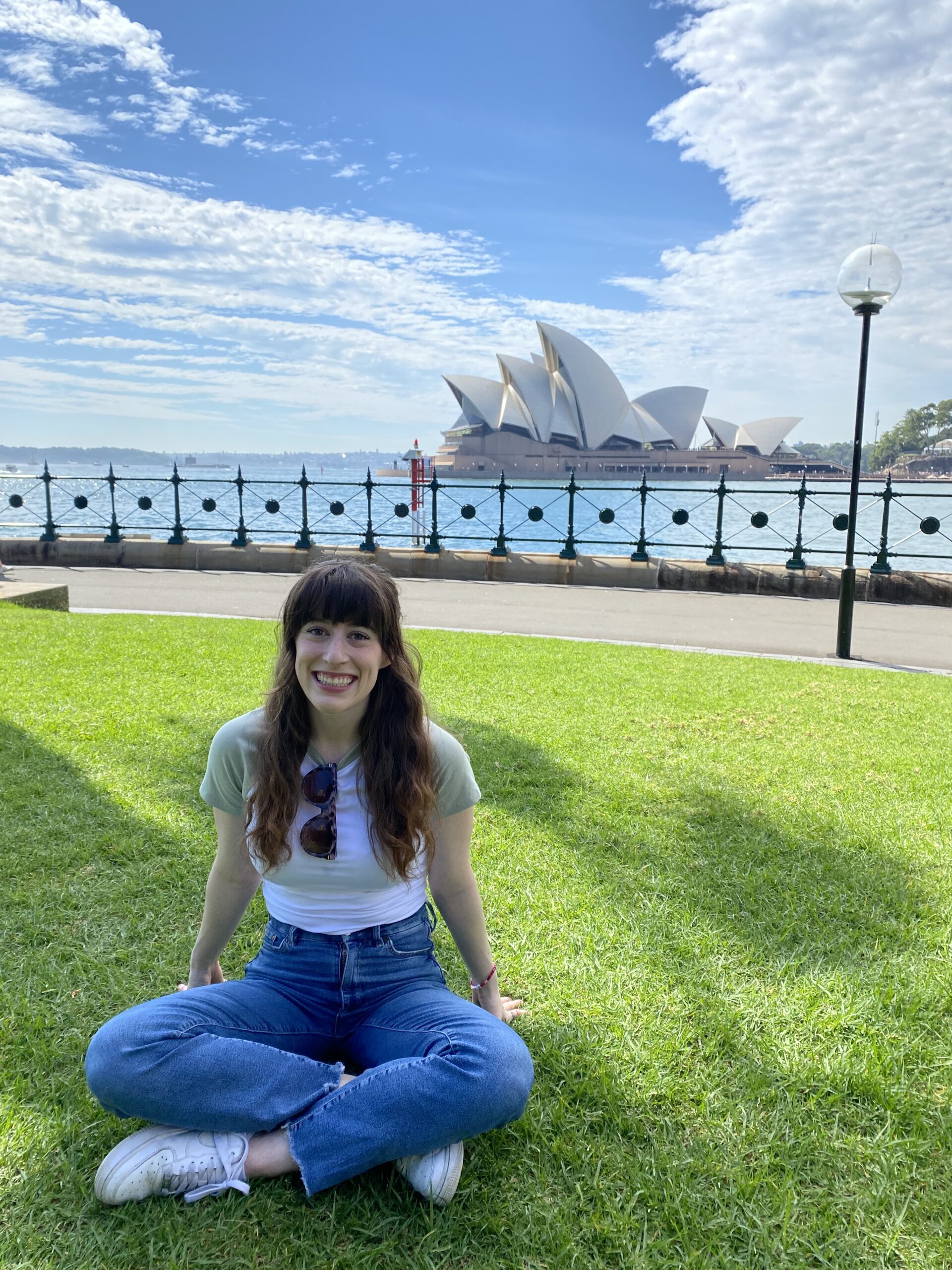 A young woman with brunt hair and bangs sitting on a grassy lawn with a view of the Sydney Opera House in the background.