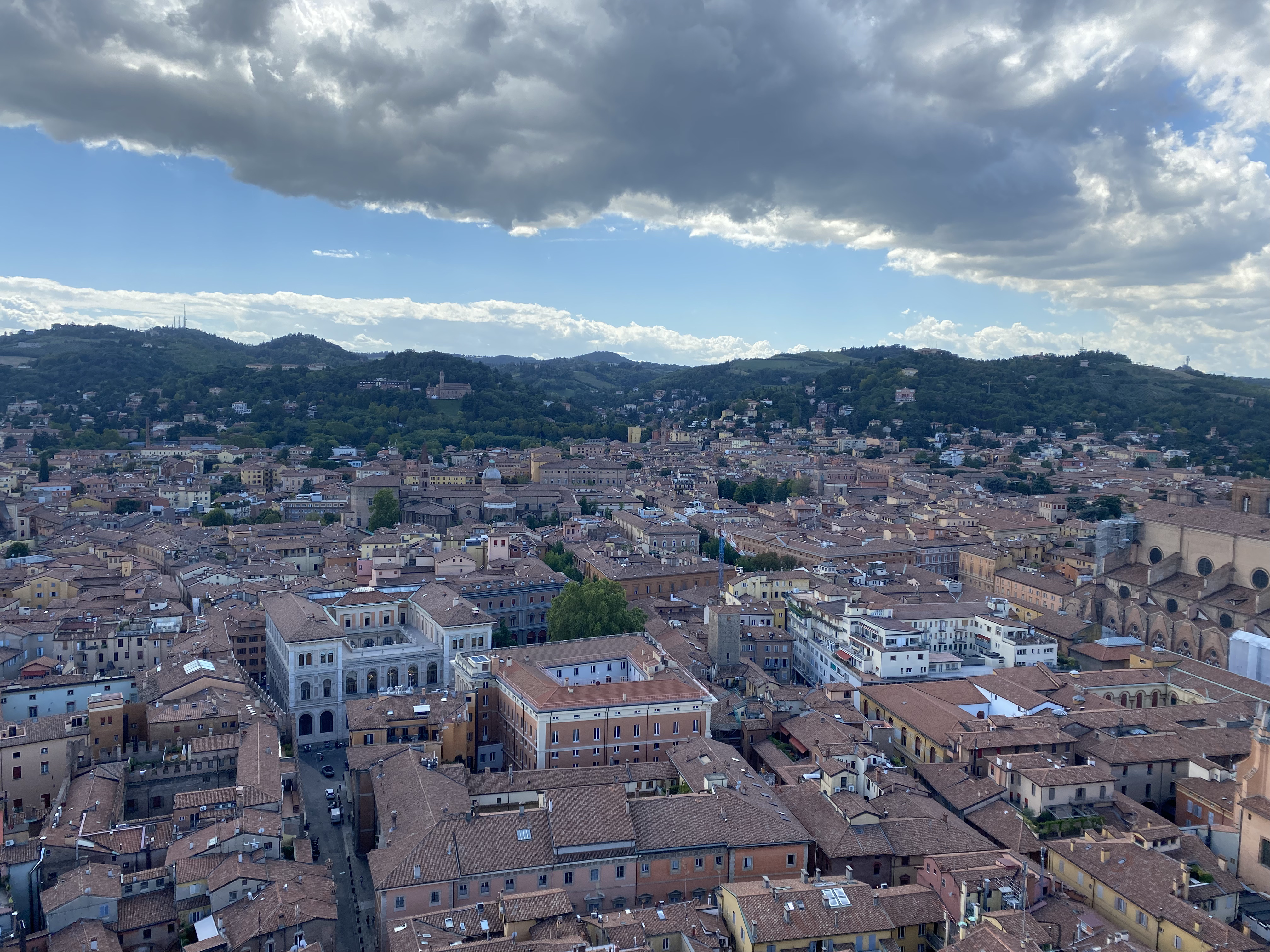 A bird's eye view of the cloud-covered city of Bologna, painted hues of orange and brown with the unique warm architecture standing out against the green landscape in the distance.
