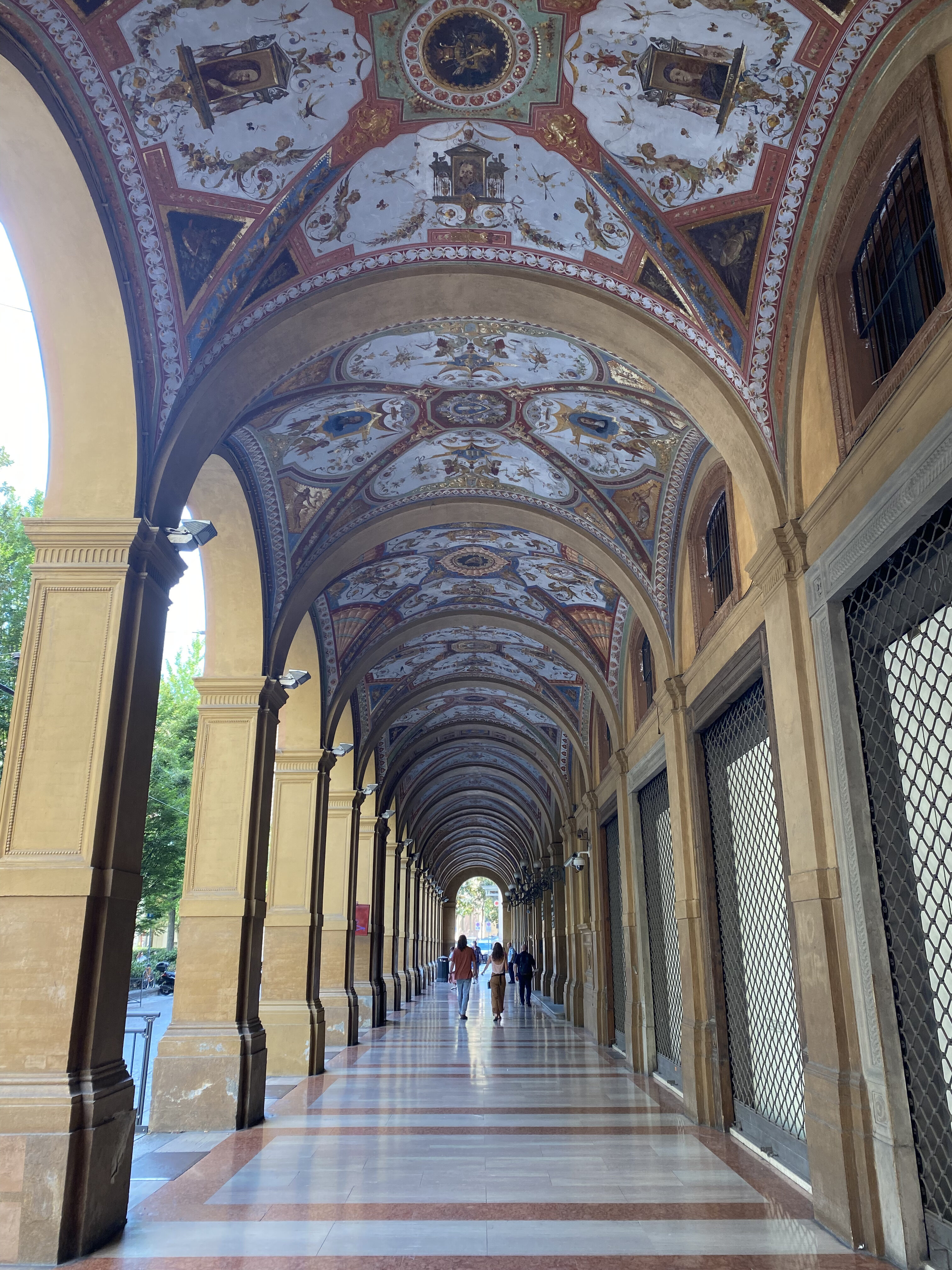 A historic portico in Bologna, Italy, with elegant arches and columns stretching into the distance, providing shade along the walkway. The warm tones of the architecture reflect the city's medieval charm and unique urban landscape.