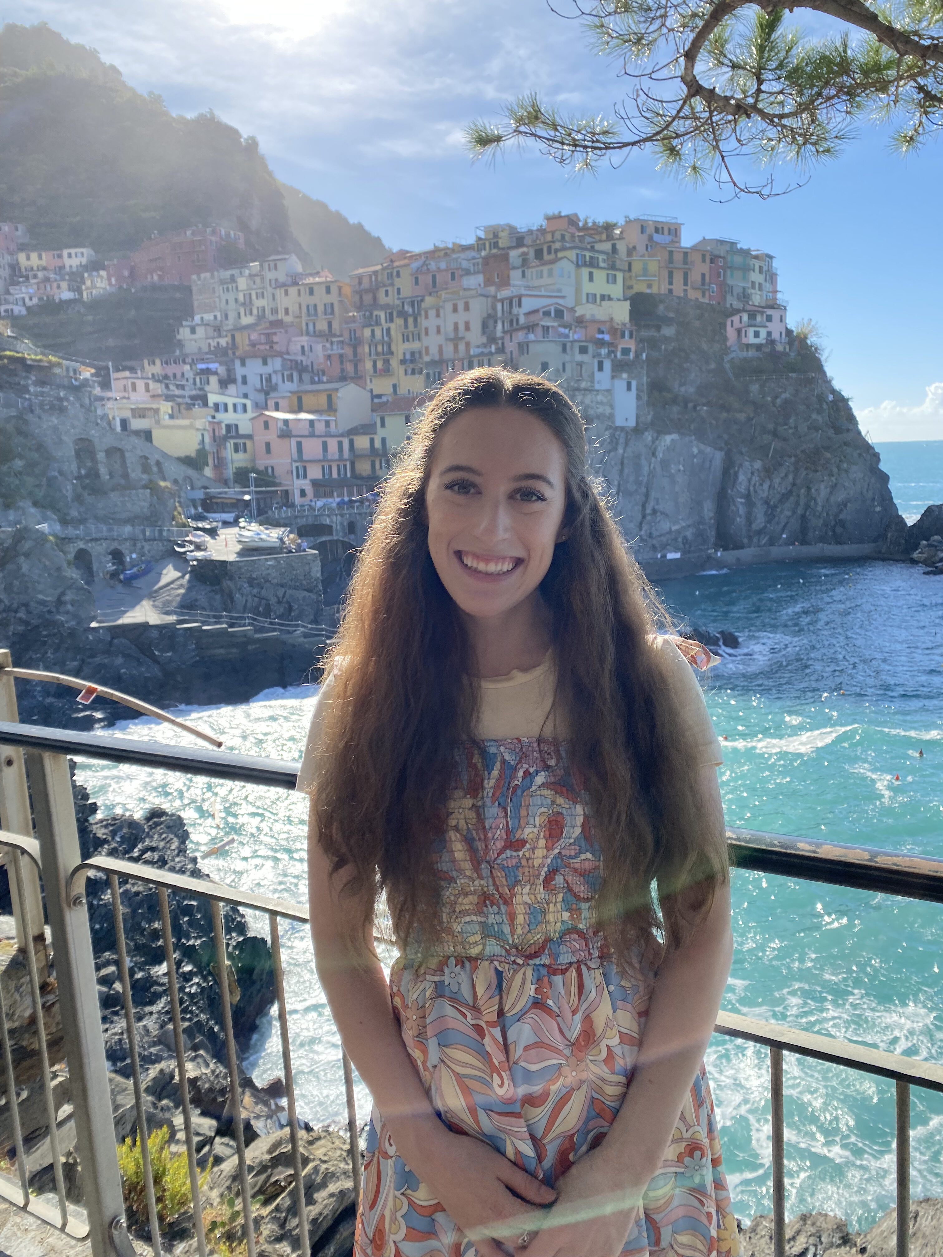 Bella, wearing a colorful dress, stands in front of a picturesque town on the Italian Riviera.