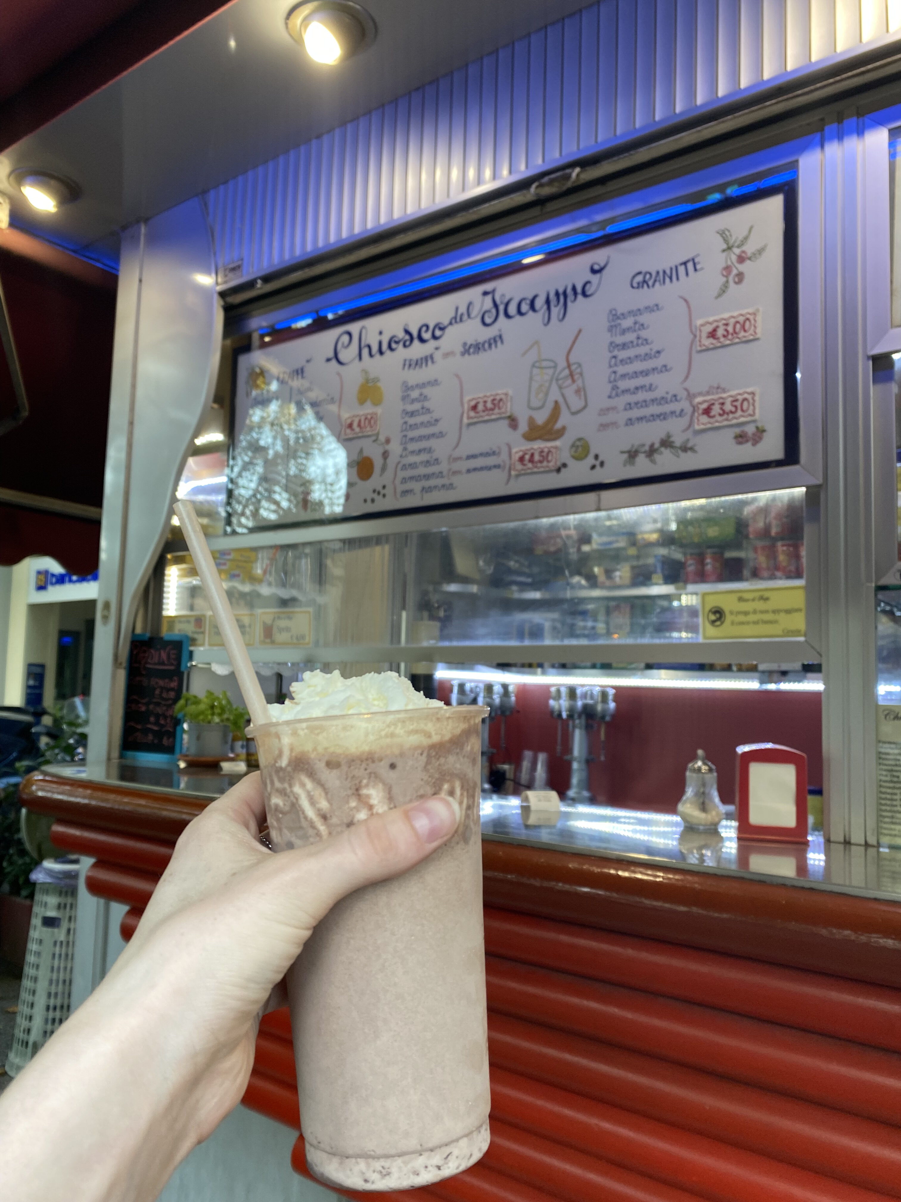 A chocolate frappe, a classic Italian milkshake, held up in front of the frappe stand from which is was purchased.