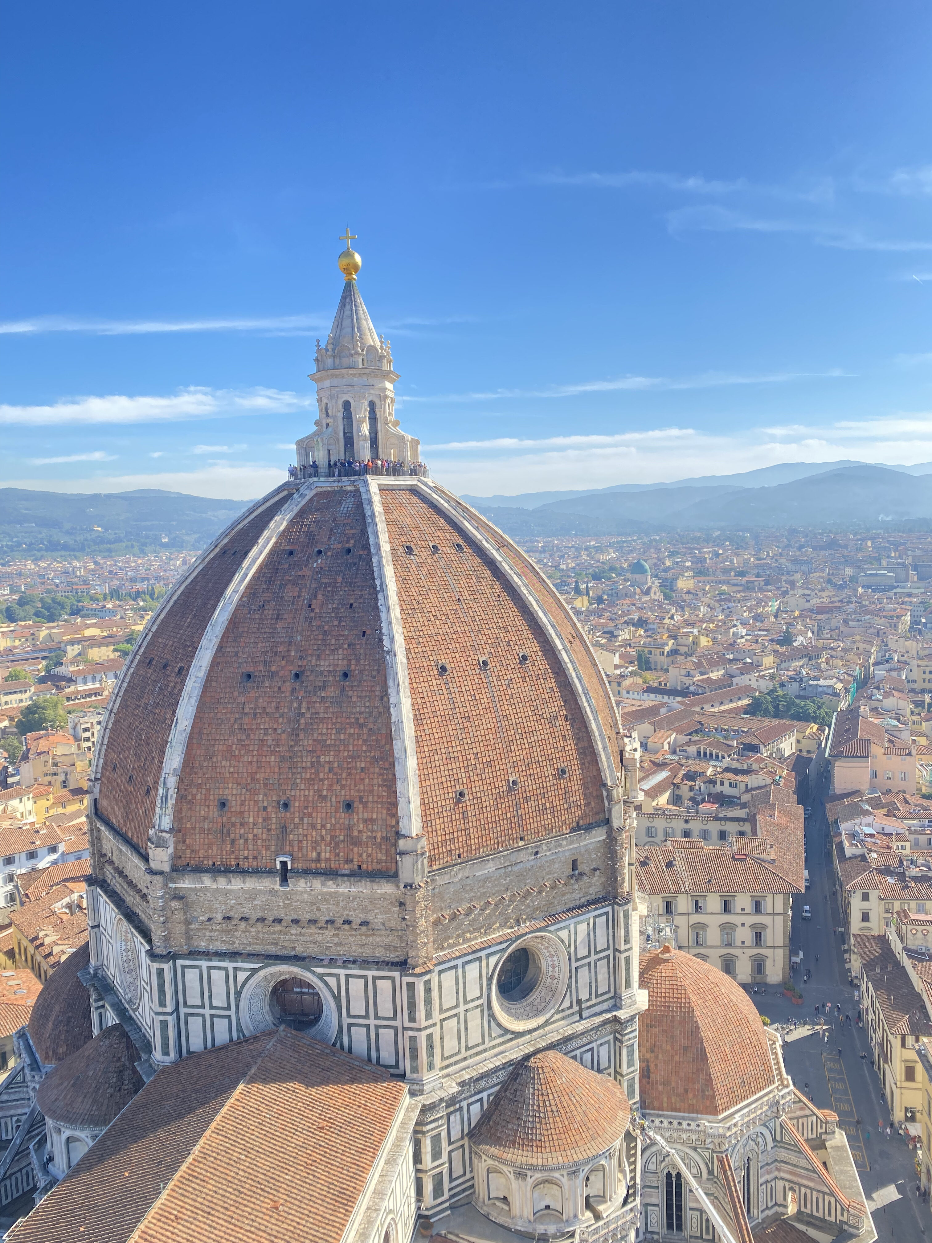 Brunelleschi's cupola in Florence, Italy, seen from the top of Giotto's Bell Tower. In the distance the orange roofs of Florentine buildings paints the city orange. The sky is bright blue.