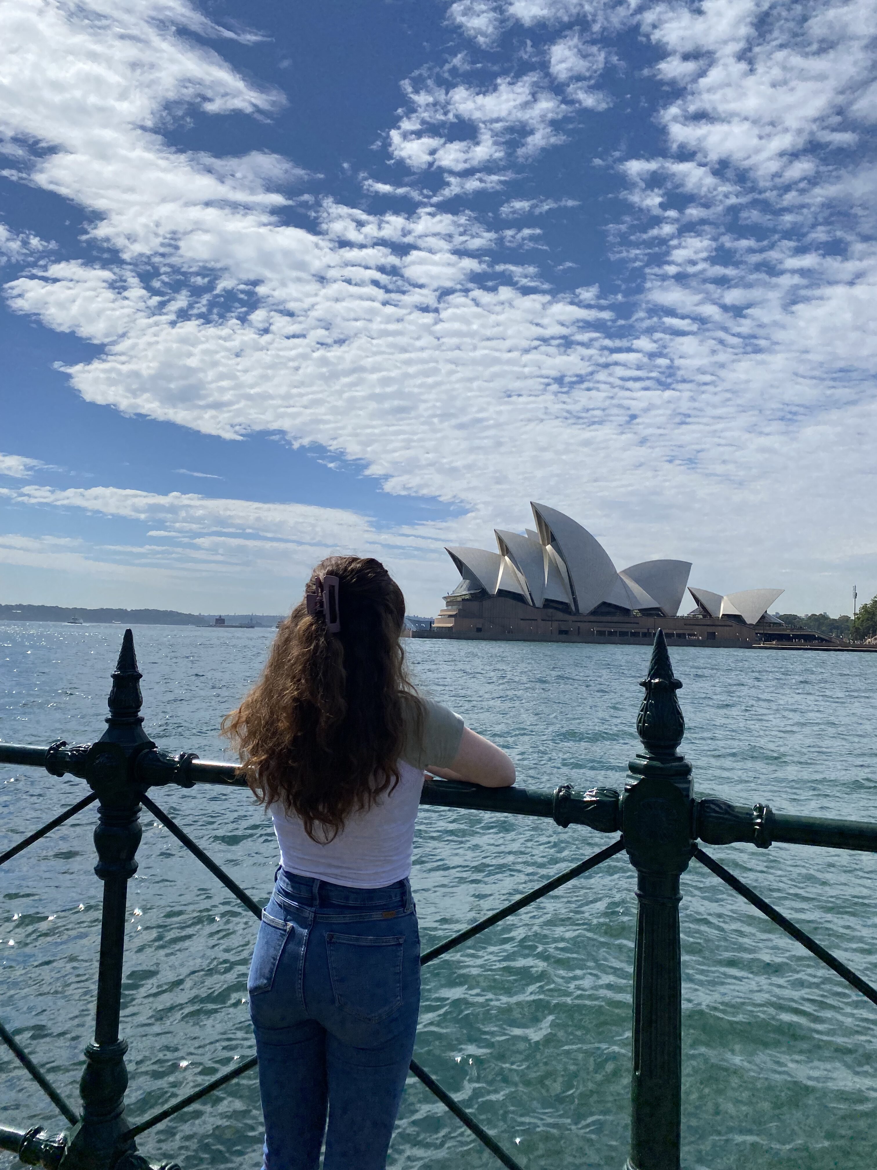 Bella looking into the distance at the monumental Sydney Opera House, leaning against a dark gate that nearly touches the harbor beneath it. A cloudy blue sky paints the horizon.