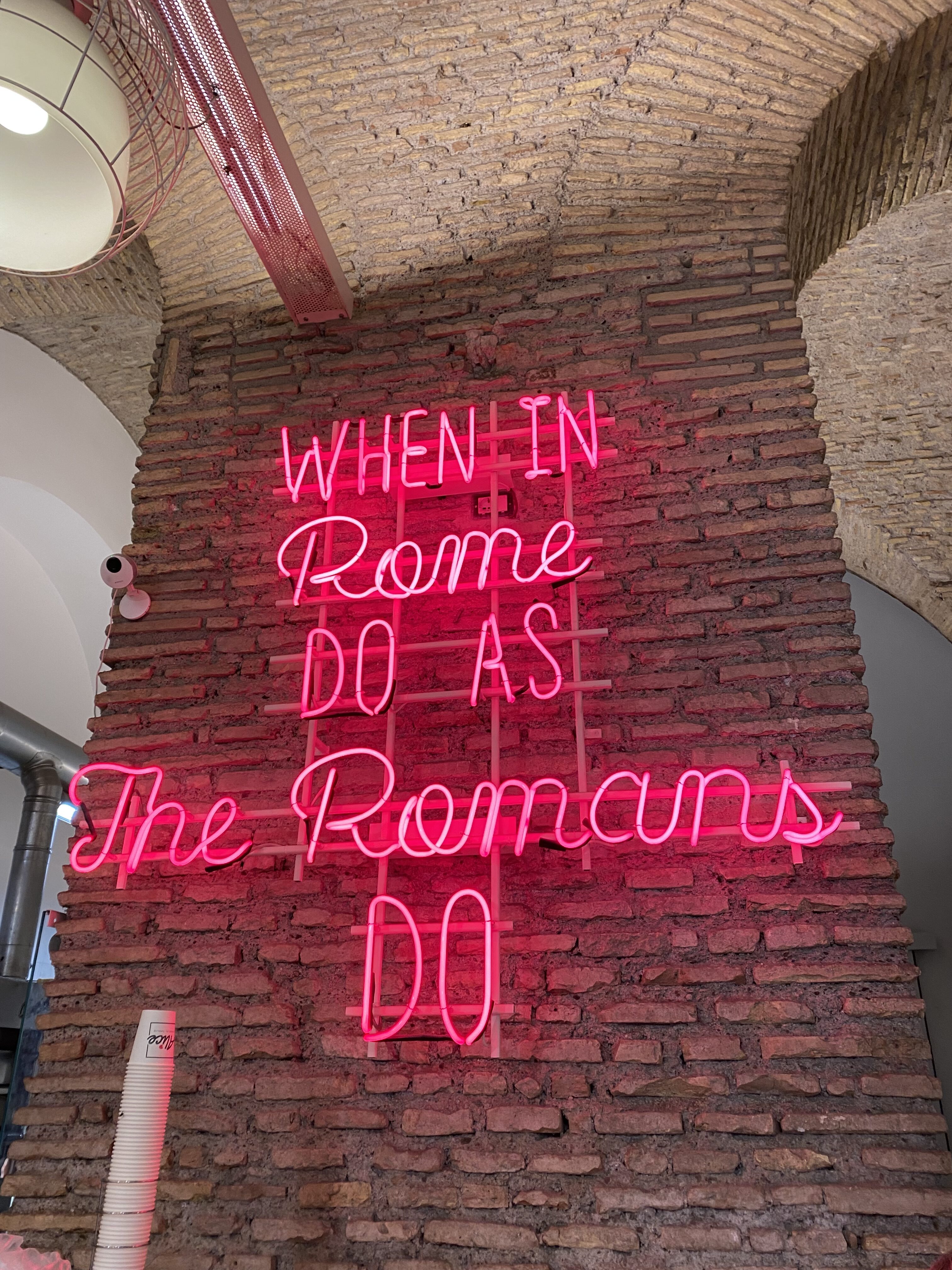 A bright, neon pink sign along a red brick wall that reads, "When in Rome, do as the Romans do".
