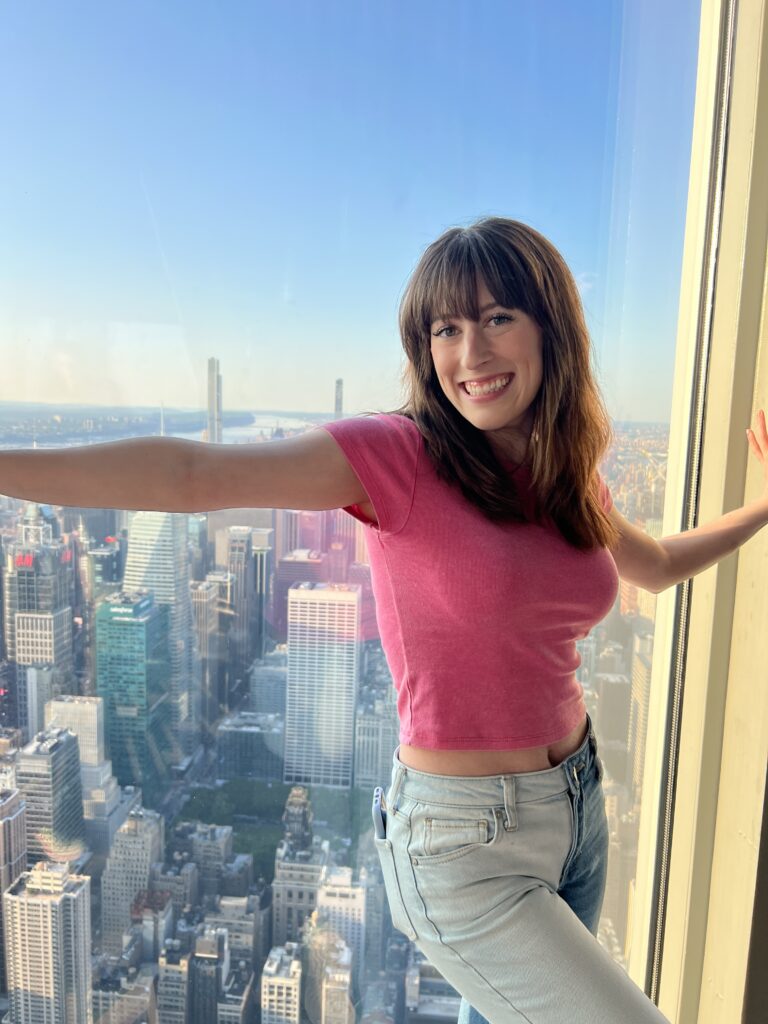 Bella posing in a pink shirt and jeans in front of the window on the Empire State Building observation deck in NYC.