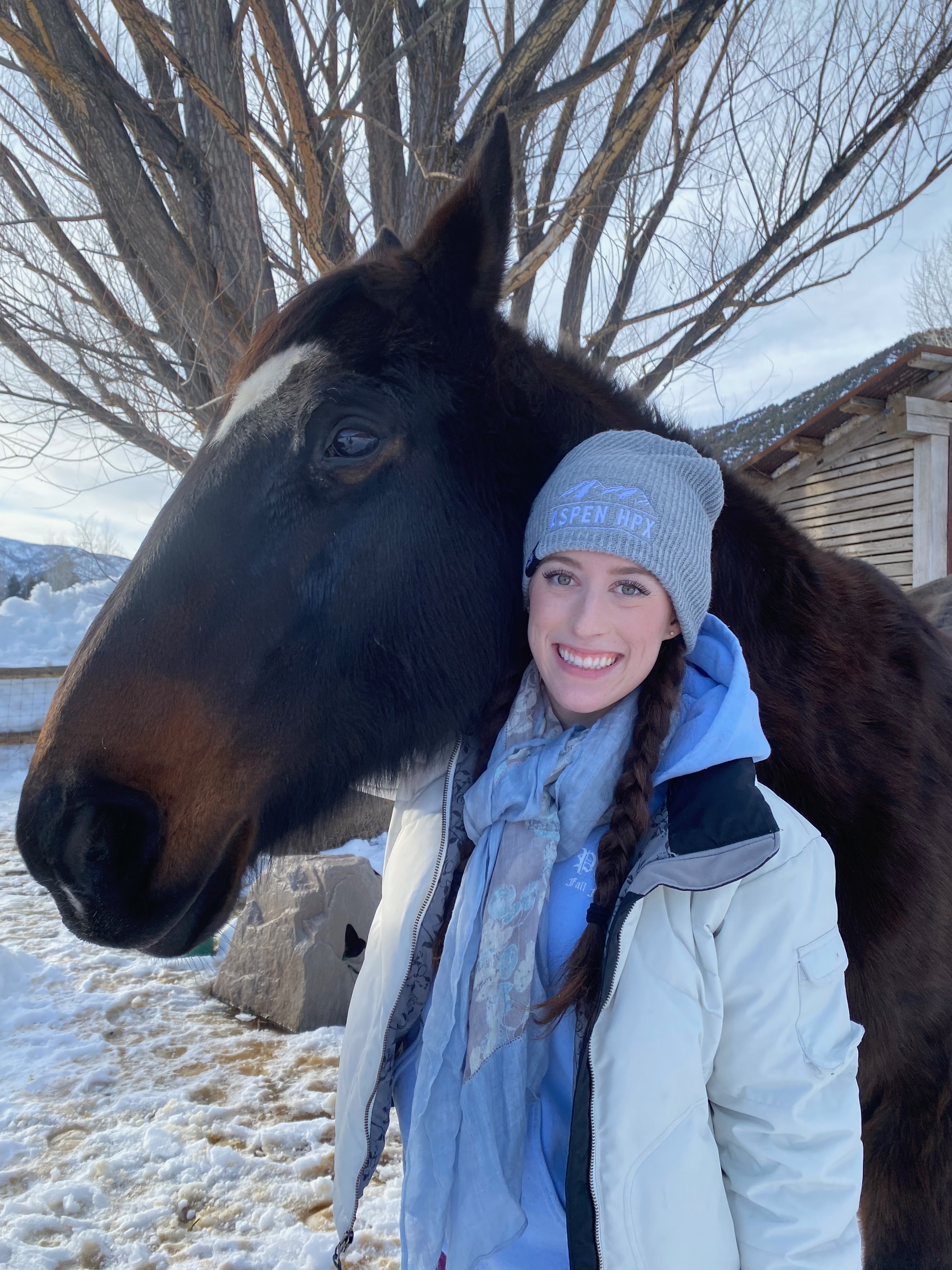 A young woman dressed in a winter coat with a beanie and braids standing beneath the chin of a beautiful dark brown horse.