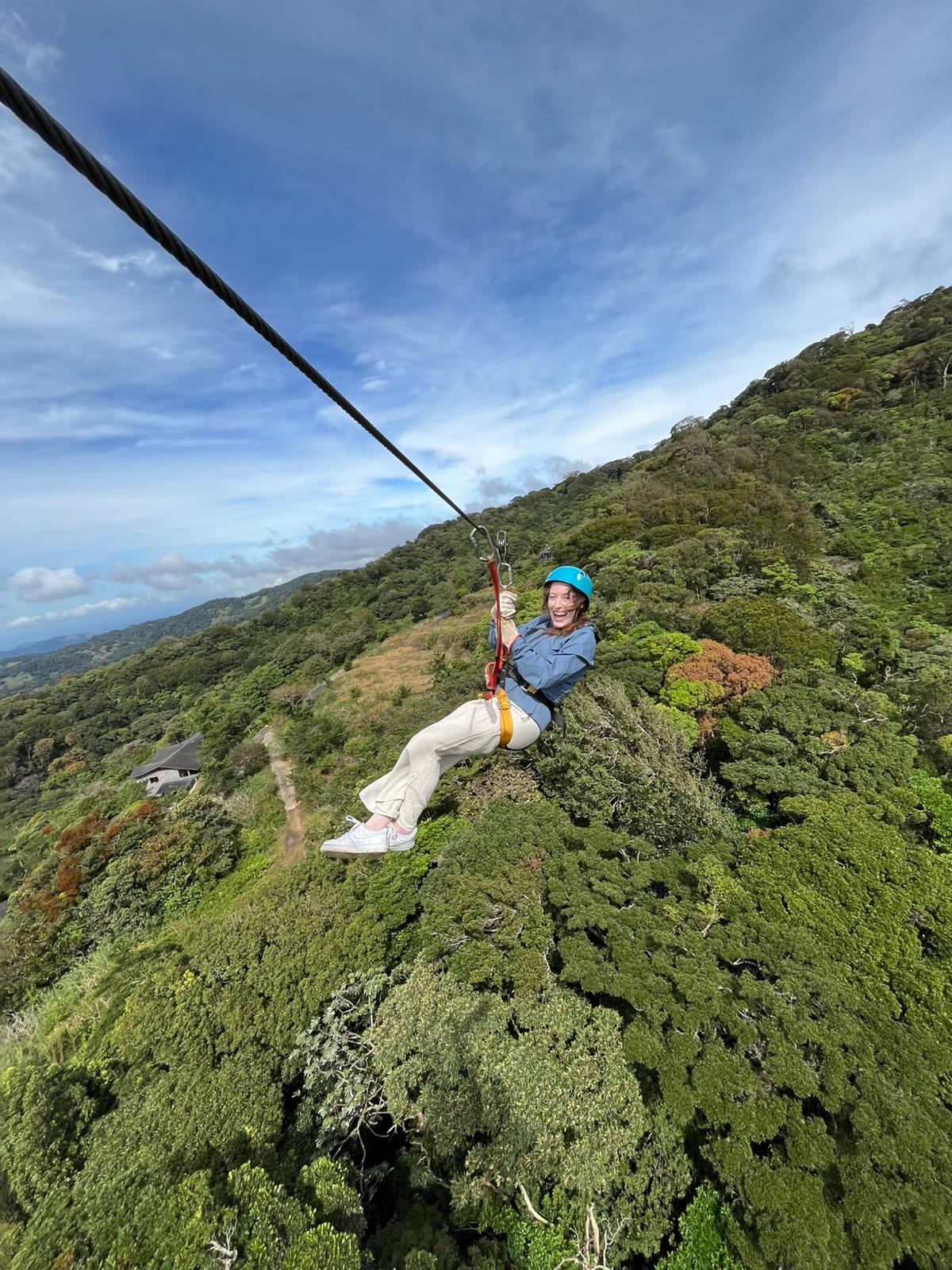 Bella smiles brightly while ziplining high above a lush, green cloud forest. She’s wearing a blue helmet, a blue raincoat, and light pants, with her feet dangling freely over the vibrant treetops. The sky is a brilliant blue with wispy clouds, and the mountains stretch out in the background — a thrilling moment captured mid-air.