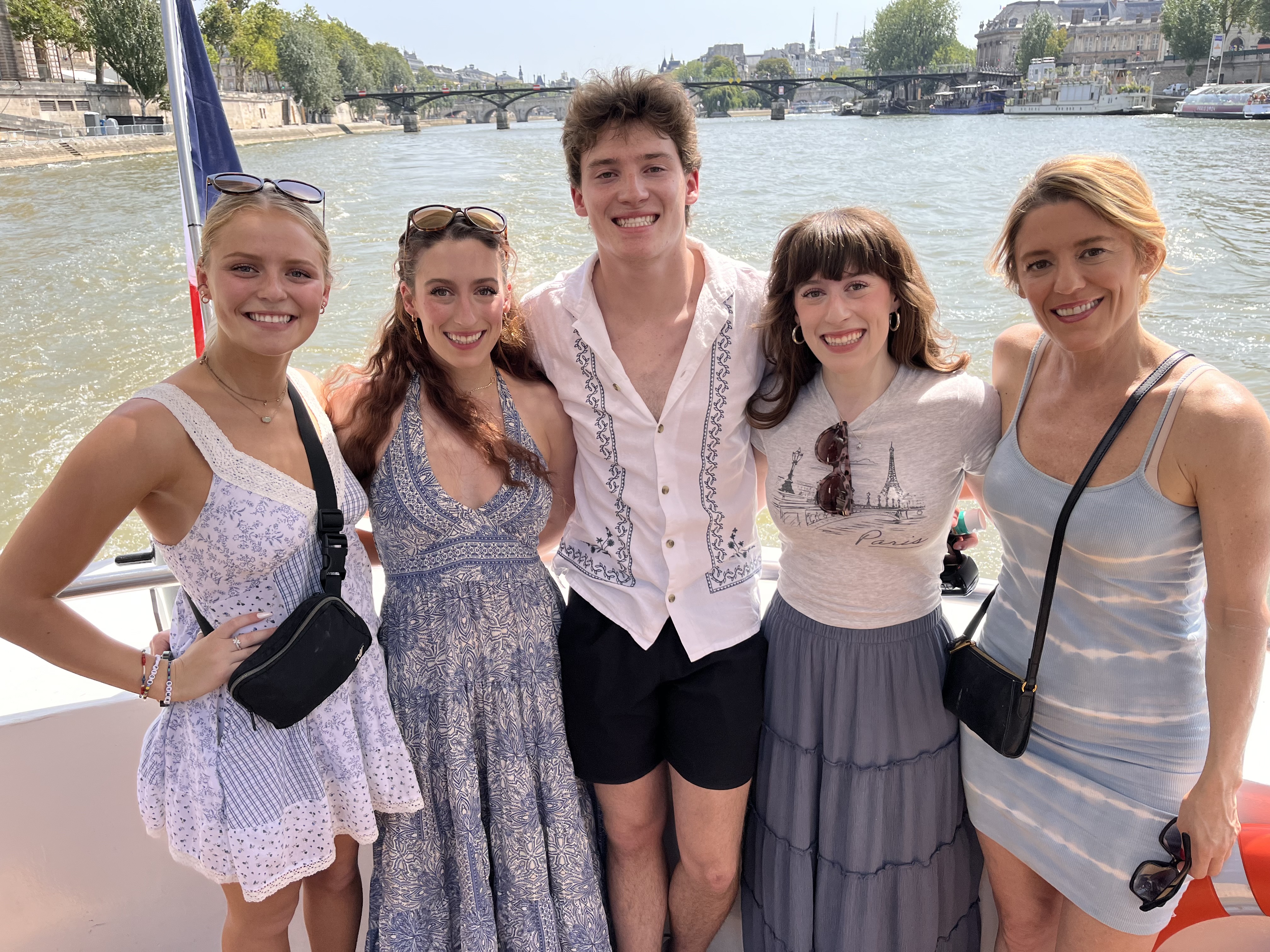 Bella and her family posing together on board a river cruise, with a French flag and the Seine river behind them. From left to right, Bella's sister Sophia, her sister Lily, her brother Liam, herself, and her mom, Cece.
