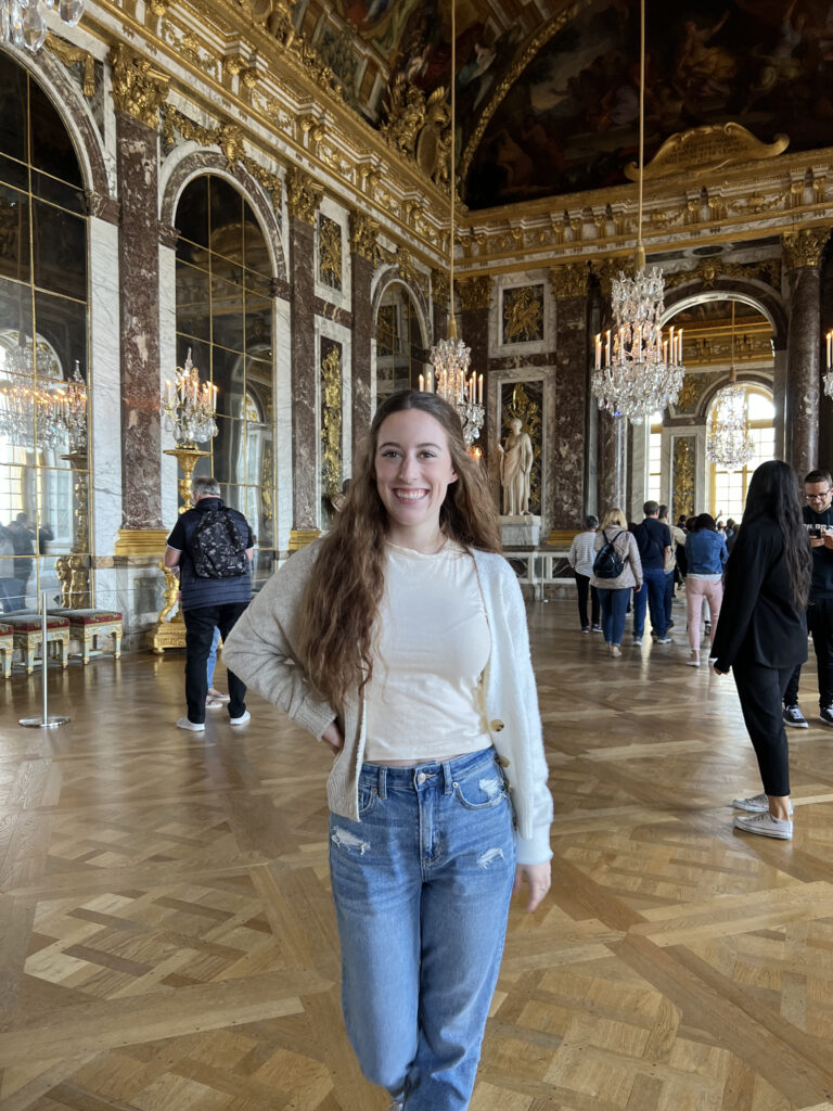 Bella, wearing a cardigan and jeans, poses in the ornate Hall of Mirrors at Versailles.