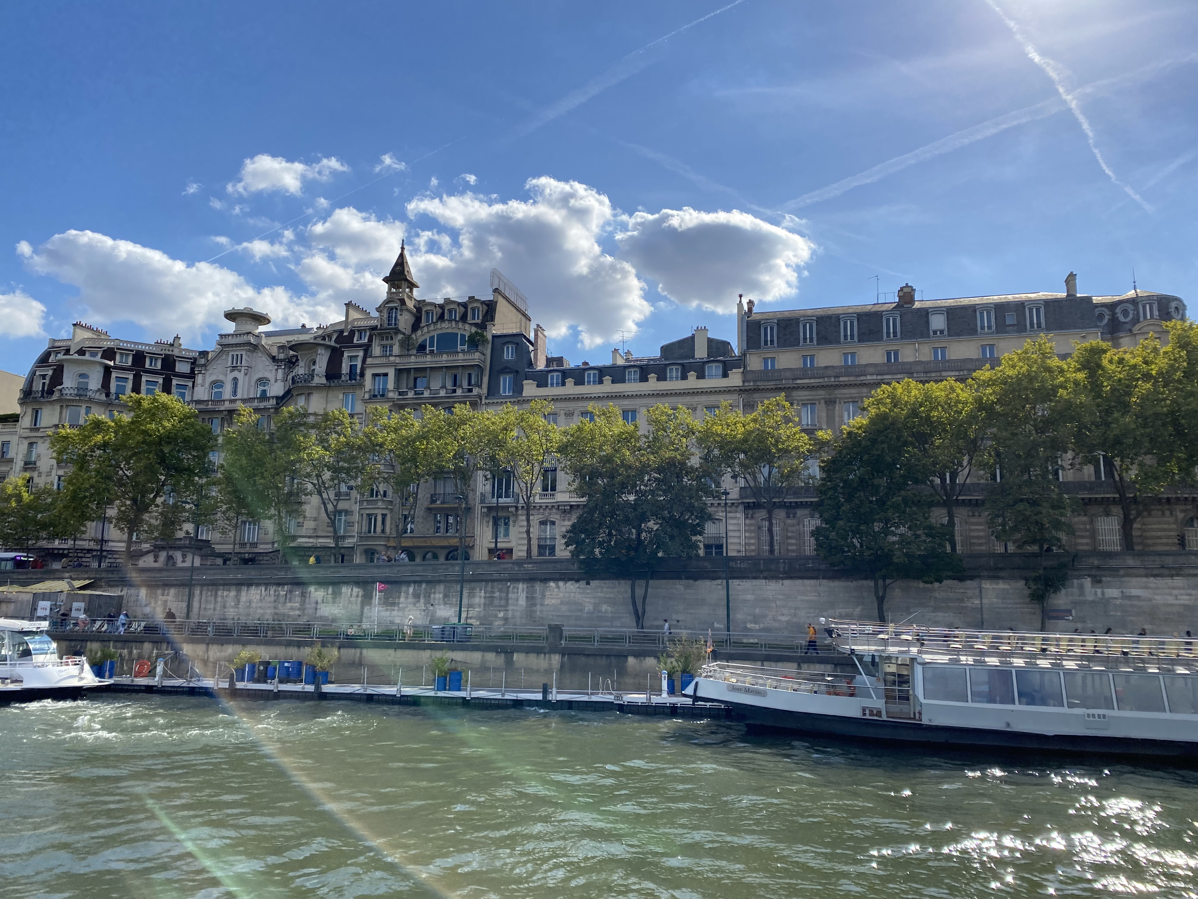 A picturesque view of the Seine river, with distinct Parisian architecture behind an array of vibrant green trees. On the river, a boat is moored. This Paris city guide features several stunning Parisian scenes.