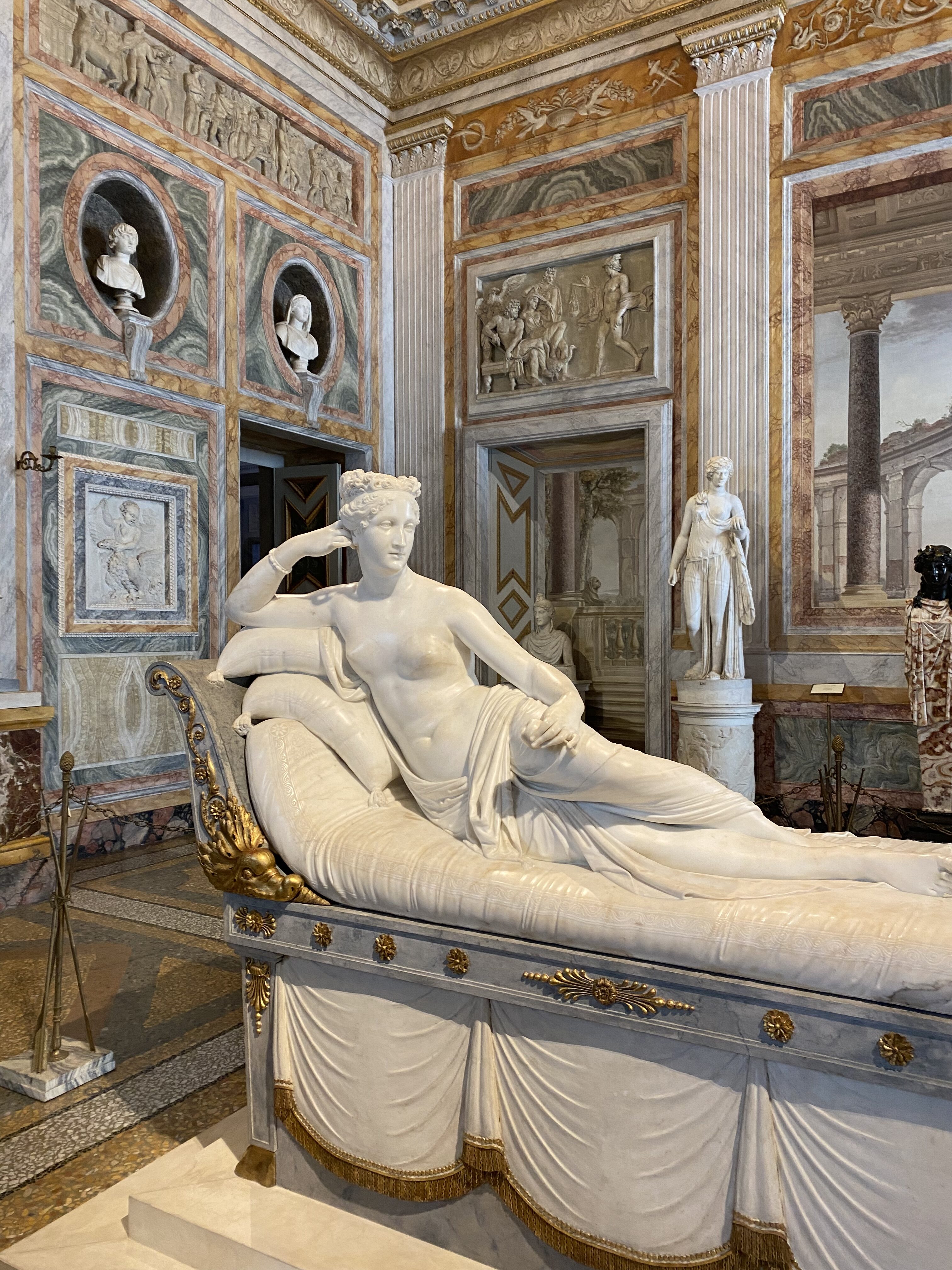 A white marble statue of Venus, reclining on a chaise lounge. The decorate marble walls of the Borghese Gallery paint the background of the sculpture.