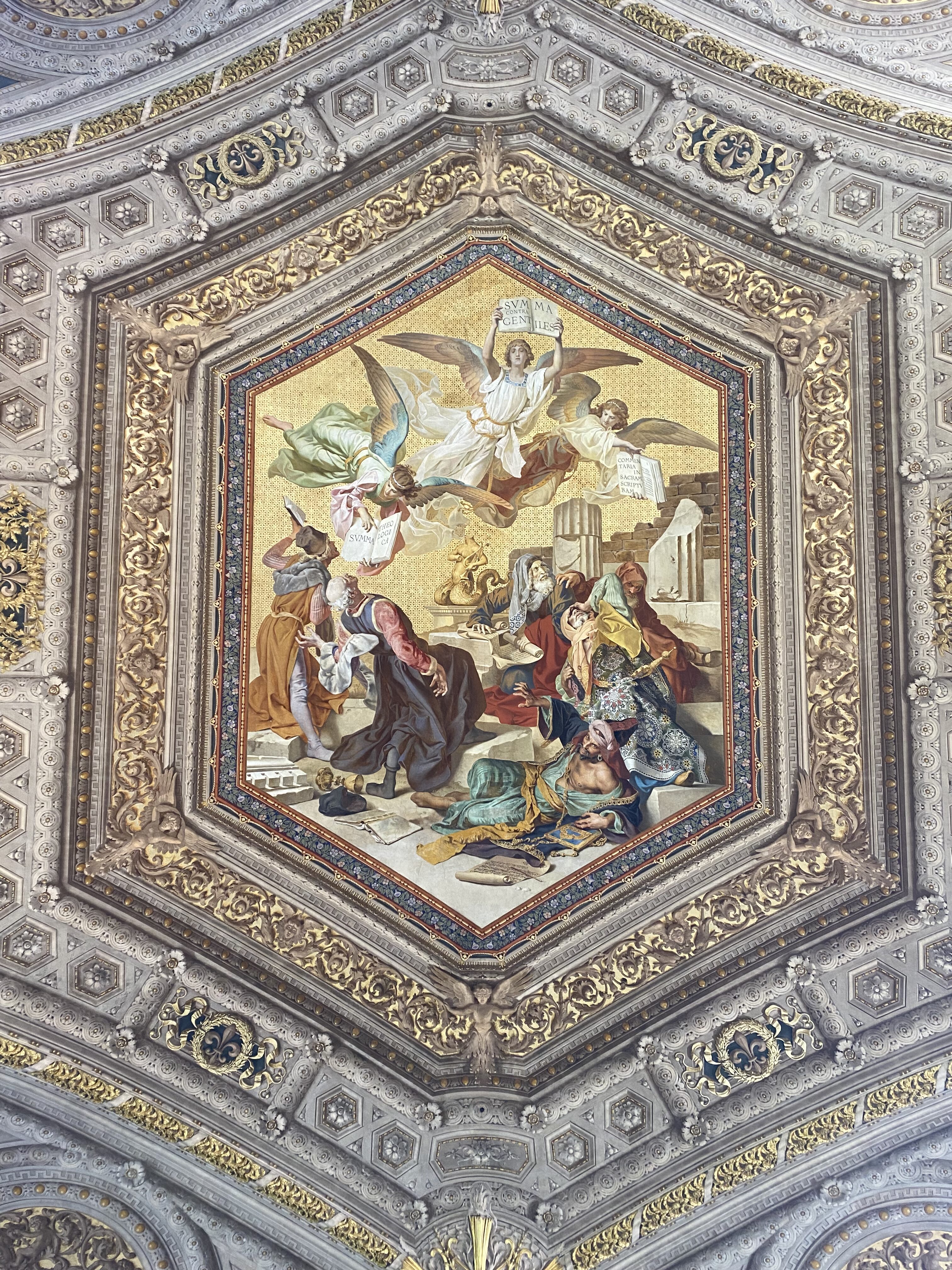 One of the ornate ceilings in the Vatican Museums. An ornately decorated ceiling in the Vatican Museum depicting angels flying above other figures, flanked by golden geometric patterns.