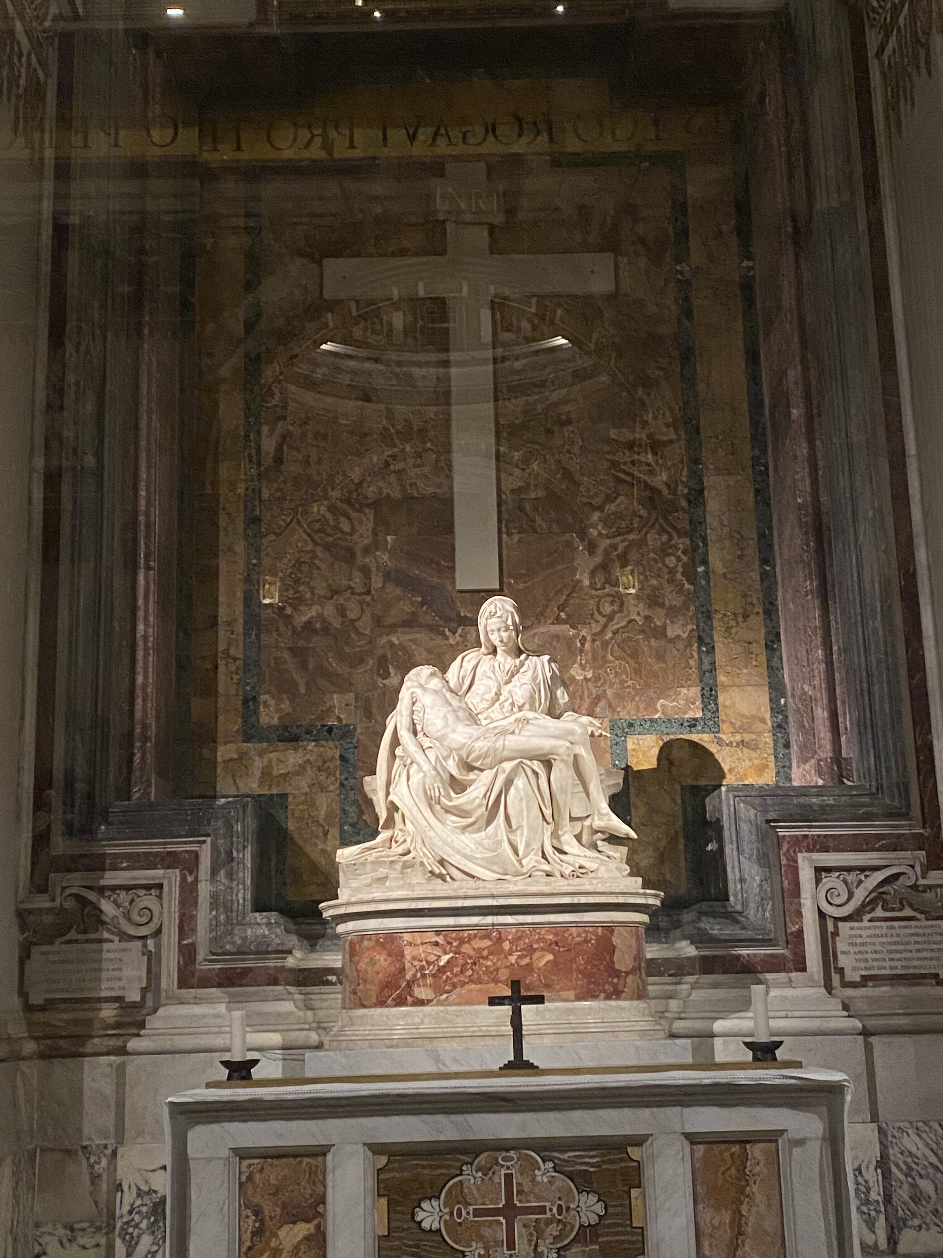 Michelangelo's famous La Pietà. A beautiful sculpture of a woman (the Blessed Virgin Mary) cradling the body of Jesus Christ after his descent from the cross. The white marble sculpture sits illuminated in front of a decorative cross.