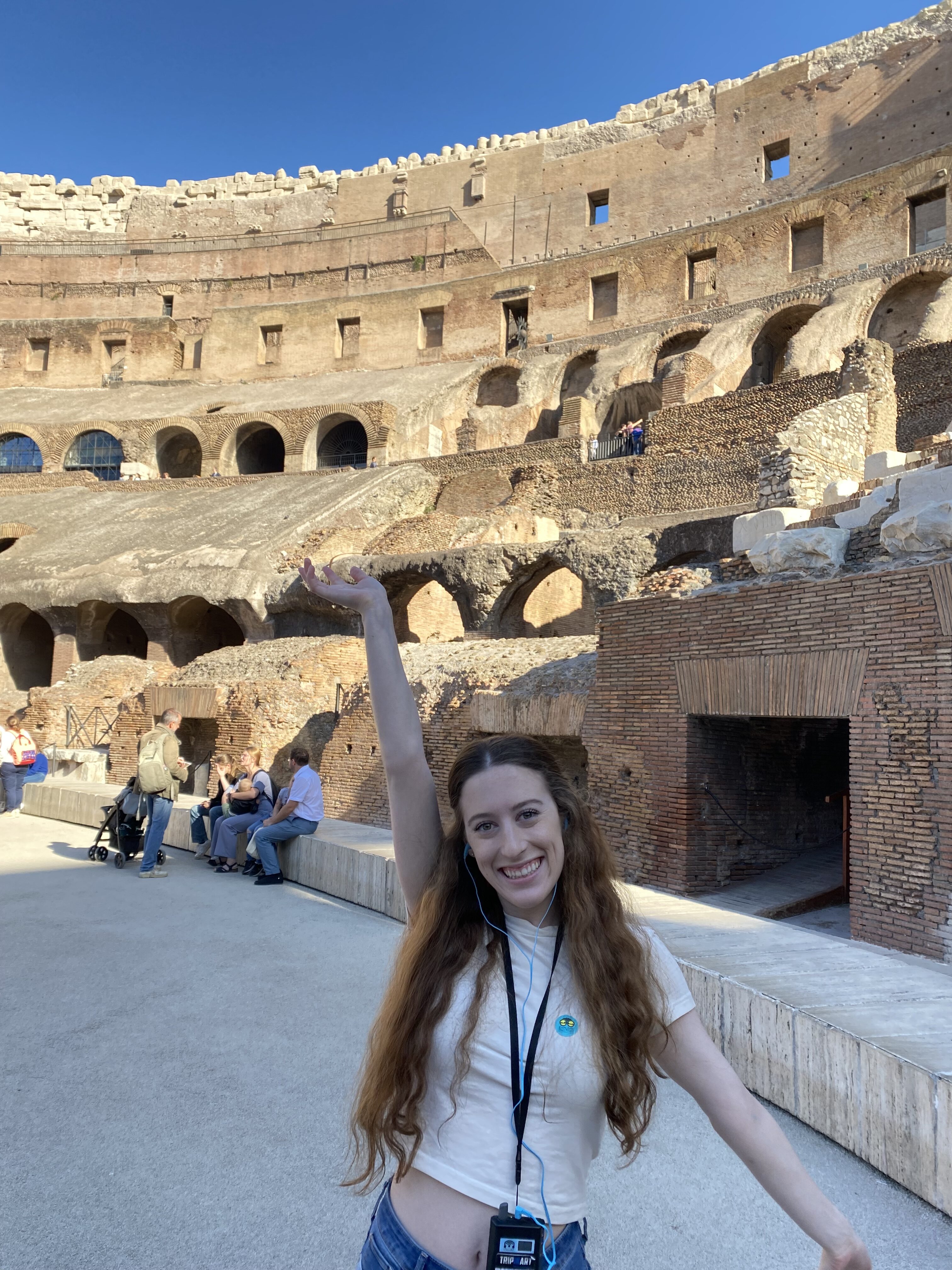 Bella poses with one arm up and the other down in the Colosseum. Around her neck is an audioguide from the guided tour.