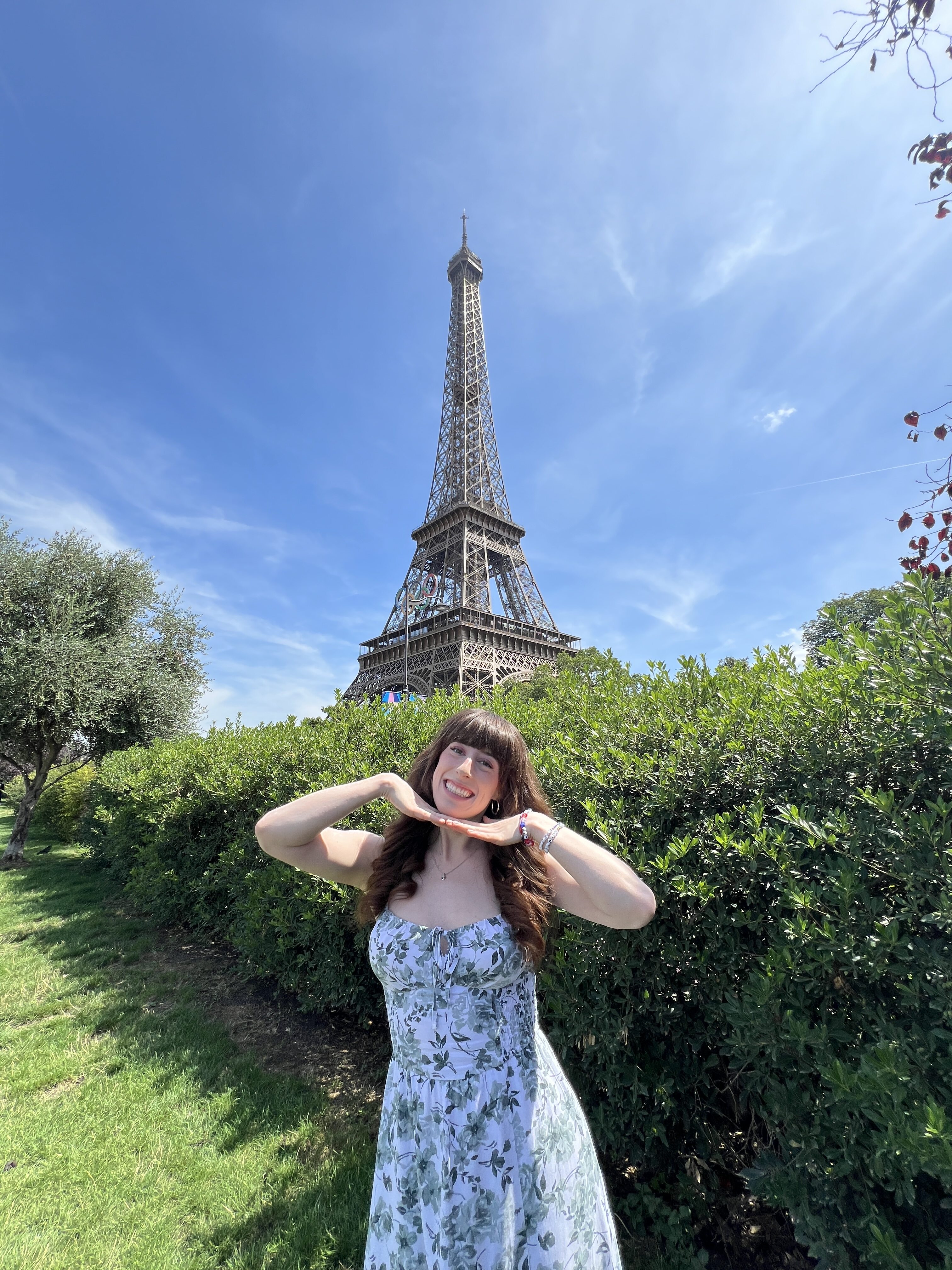Bella, in a white dress with green flowers, posing in front of a green bush with her hands beneath her chin and the Eiffel Tower in the background.