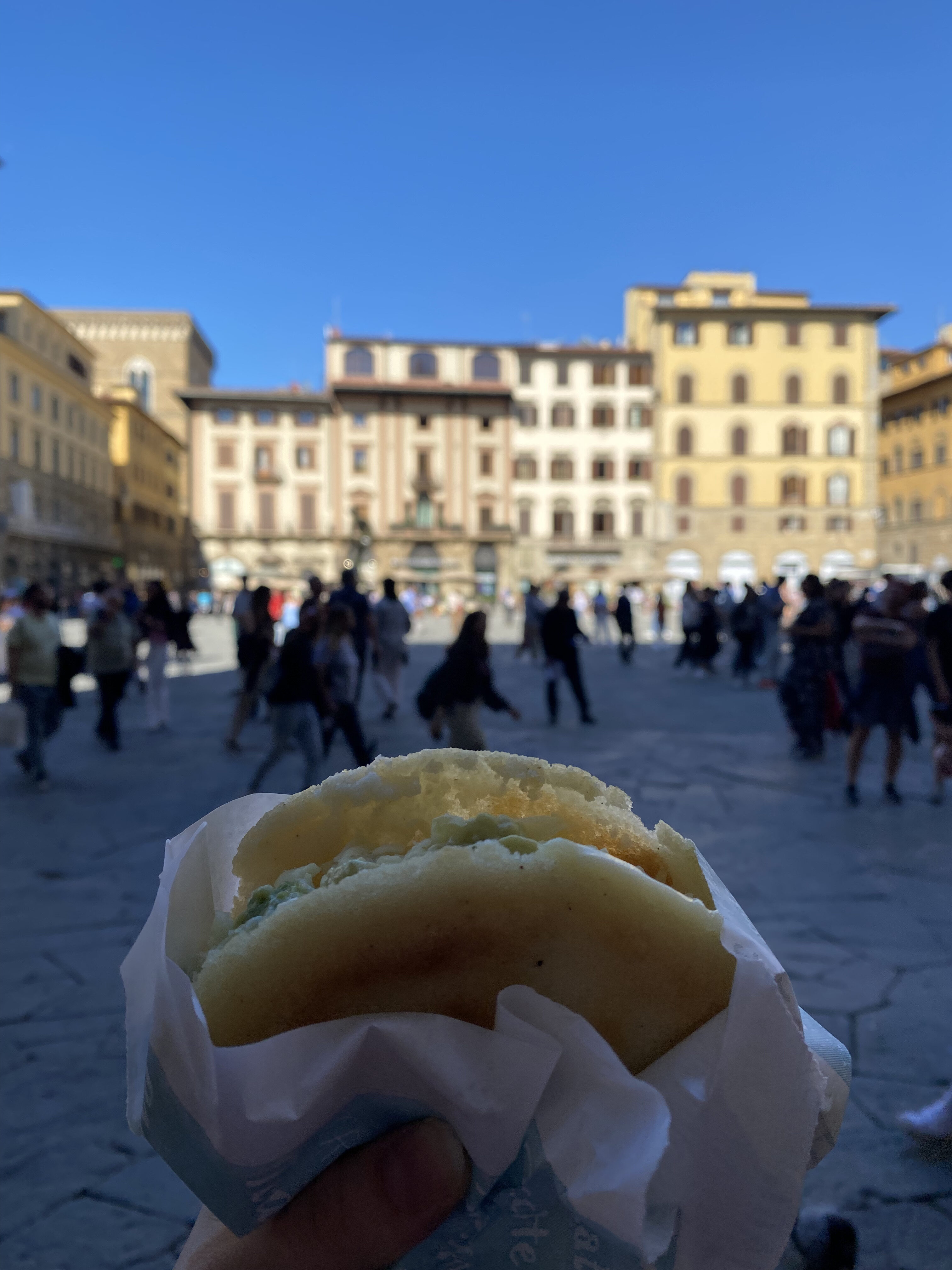 A golden arepa wrapped in white paper. In the background is a shaded piazza with illuminated Florentine buildings in the background.