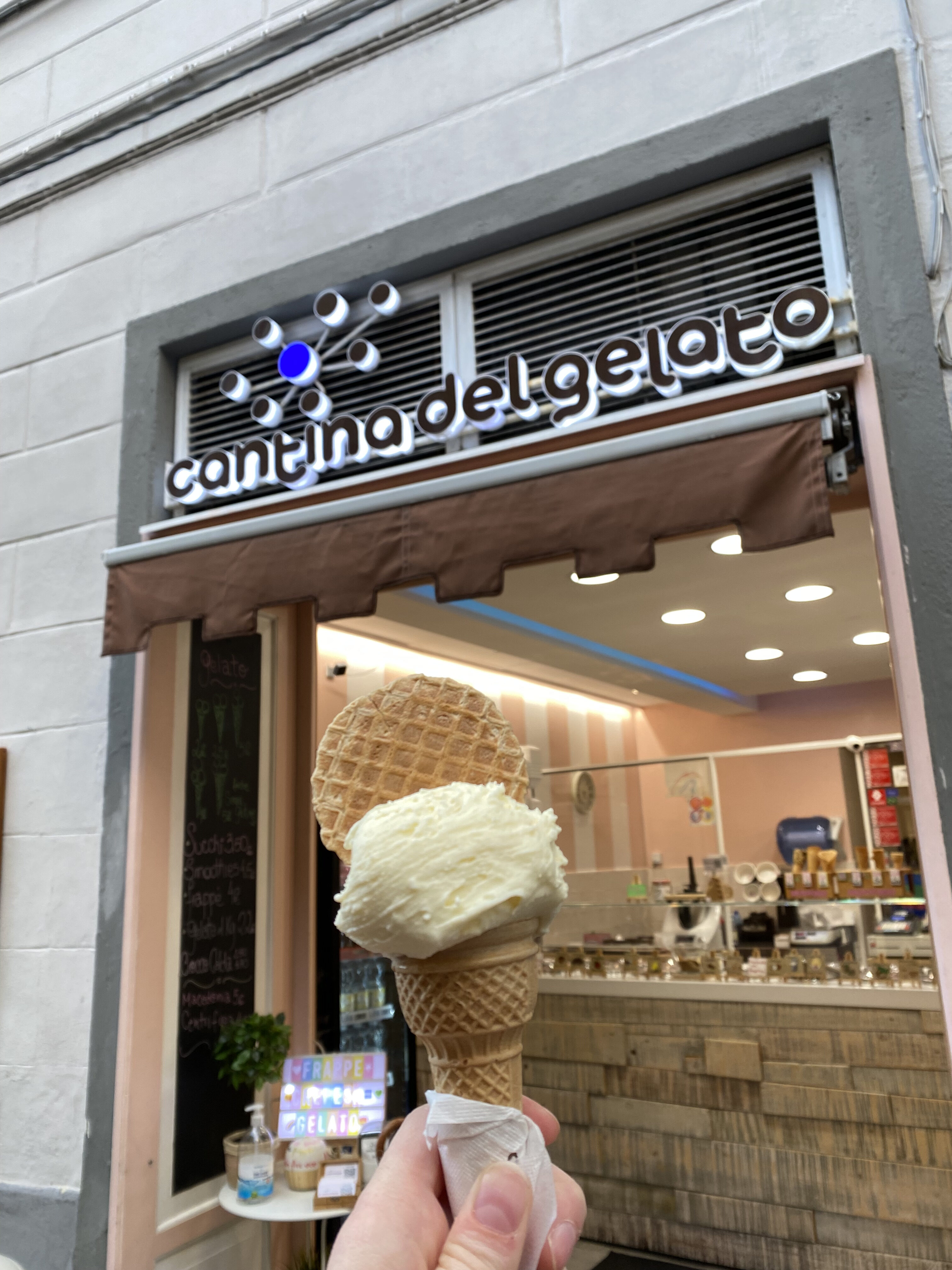 A small cone of creamy gelato held in front of a Gelateria with a sign reading, "Cantina del Gelato" in Florence, Italy.