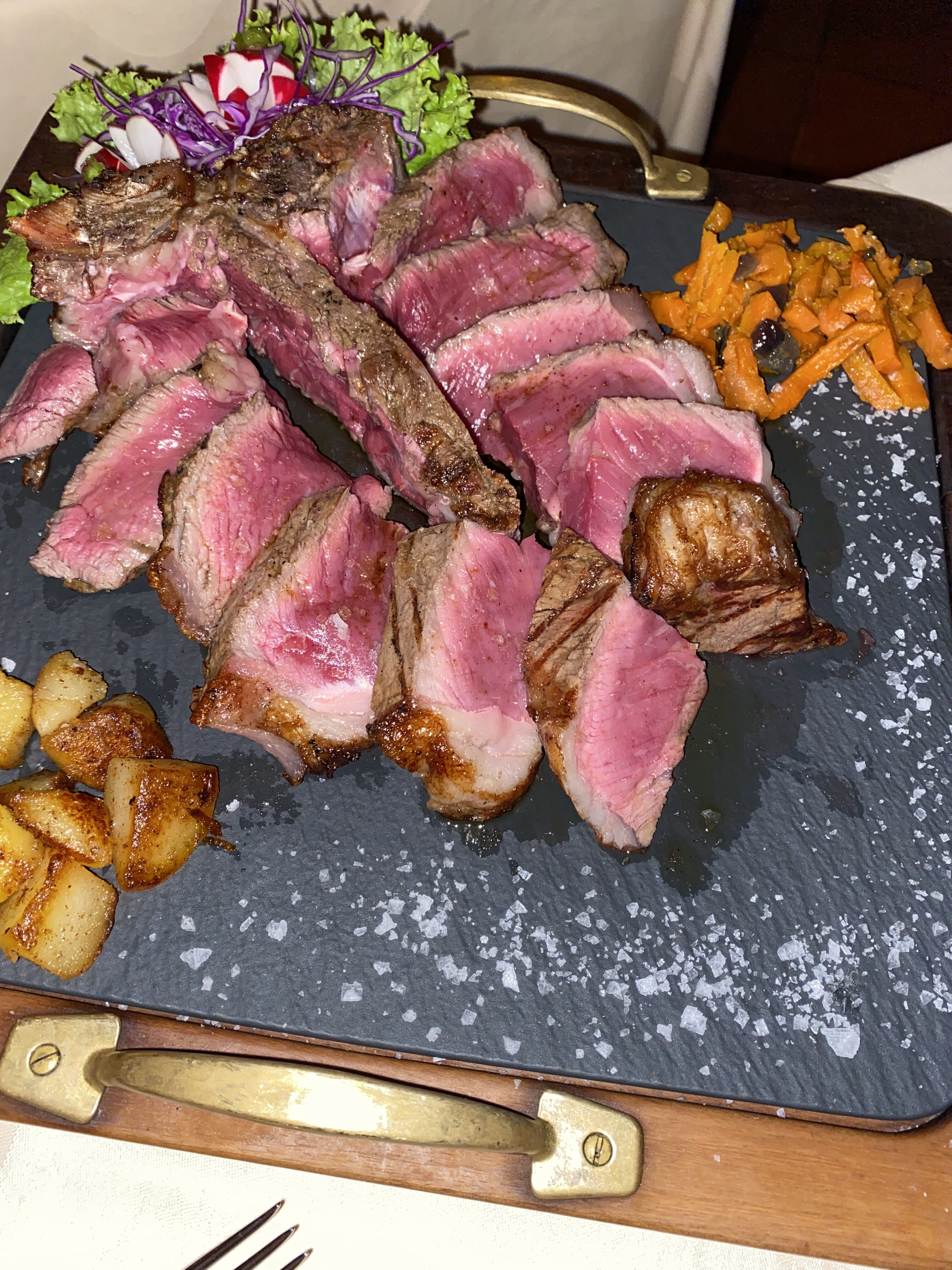 A rare Florentine steak plated with herbs and potatoes on a black skillet.