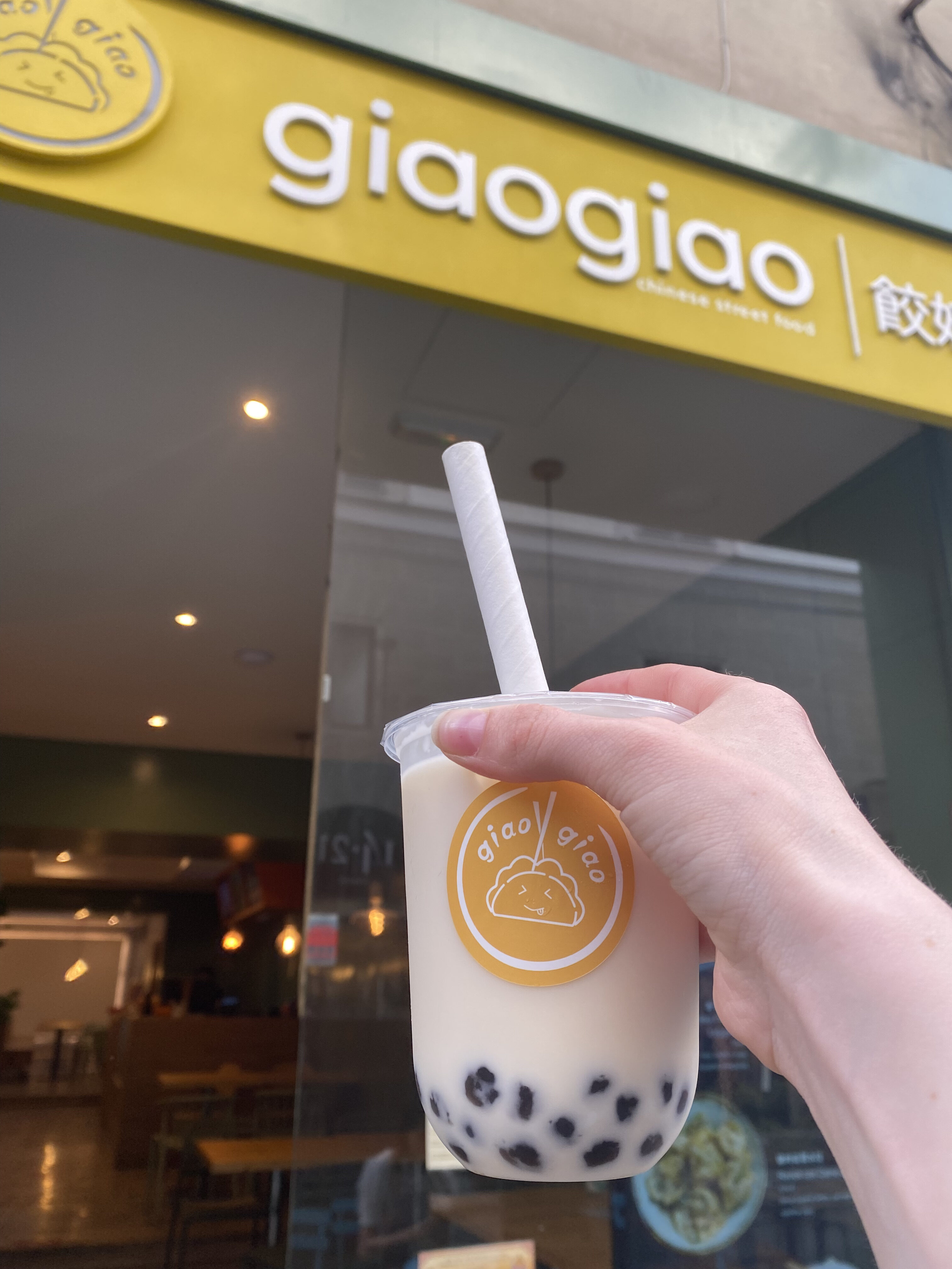 A cup of milk tea with a bright orange logo that reads, "GiaGiao". Tapioca boba pearls sit at the bottom of the cup. In the background, the restaurant sign reads, "GiaoGiao".