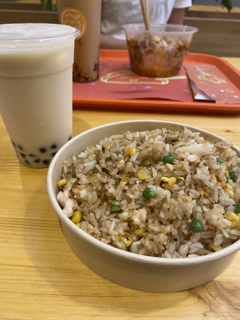 A bowl of Chinese fried rice with a cup of milk tea with tapioca pearls on a yellowish wooden table.