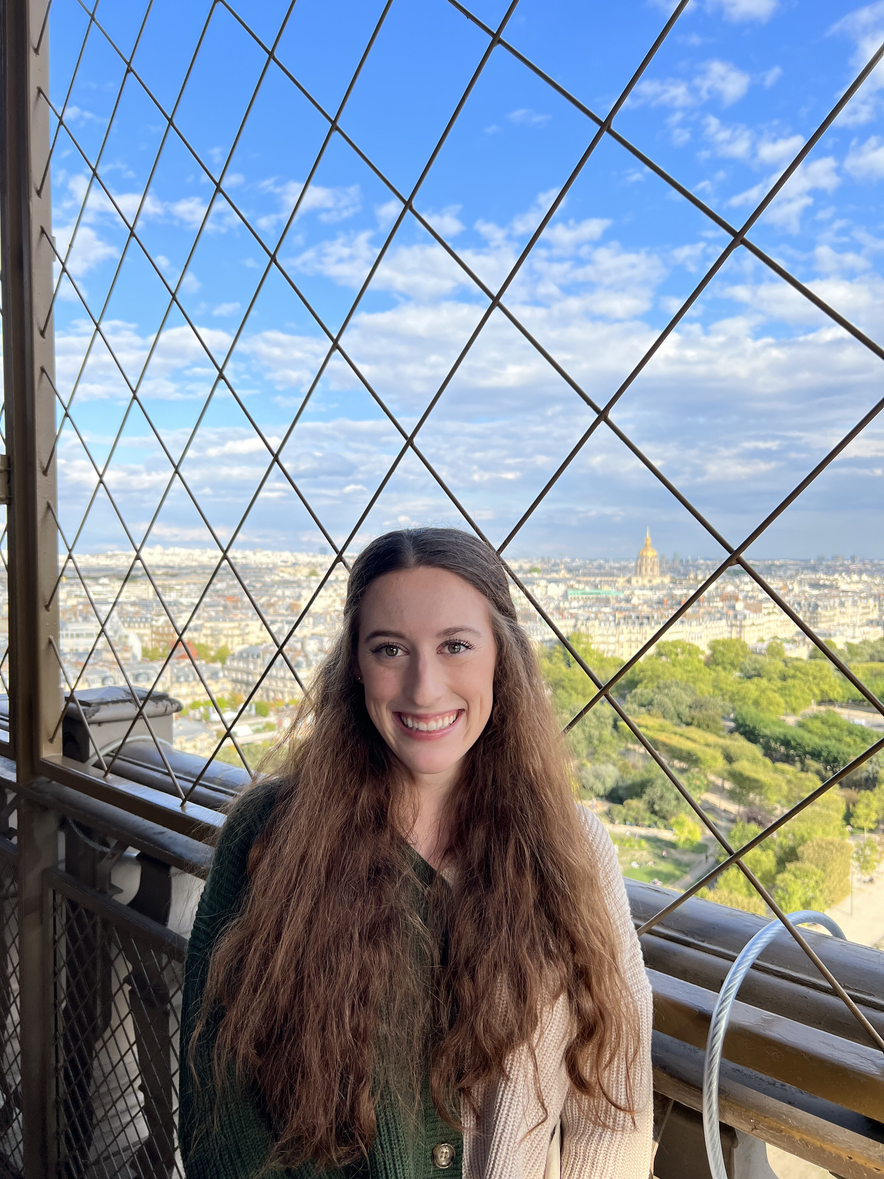 Bella poses in front of a wrought-iron lattice fence at the top of the Eiffel Tower. Through the thin fencing the city of Paris is visible.