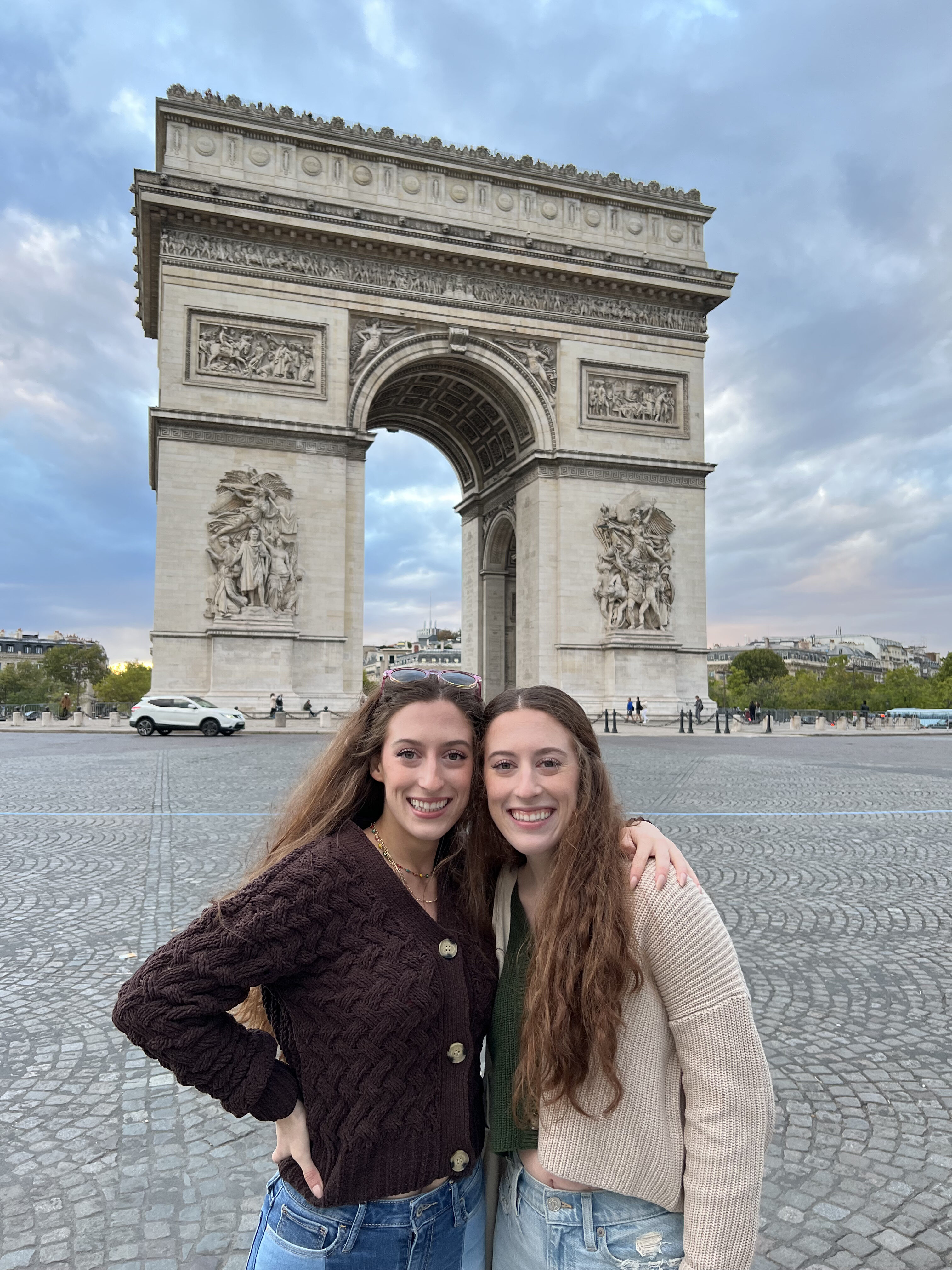 Bella and her twin sister Lily pose in front of the famous street surrounding the Arc de Triomphe. A cloudy sky is beginning to see the first signs of sunset behind them.