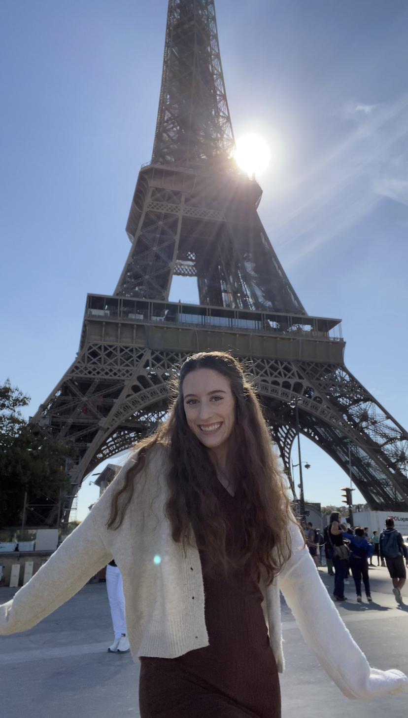 Bella wearing a brown dress and a cream sweater in front of the Eiffel Tower