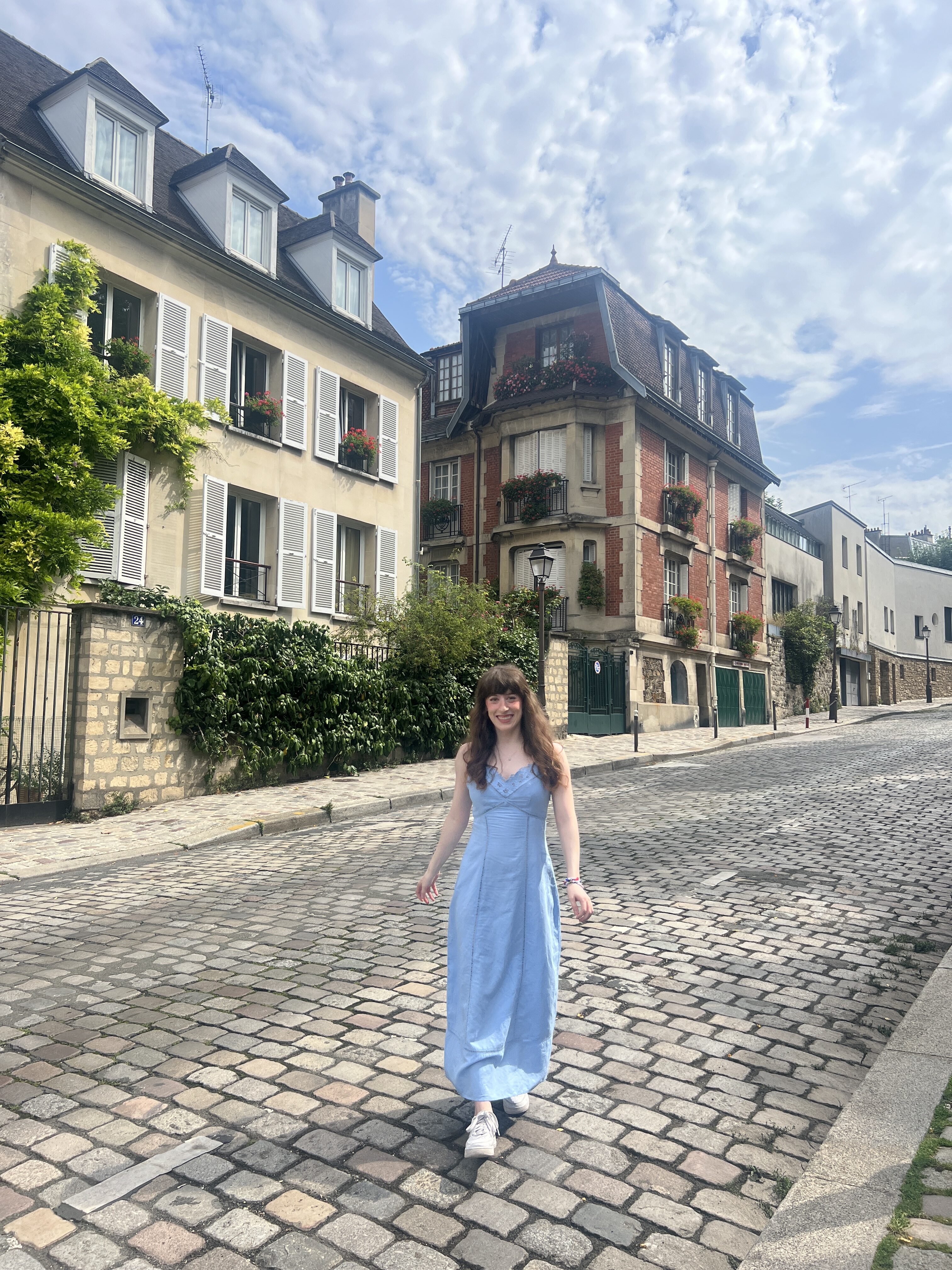 Bella wearing a sky blue dress walks along a charming street in Montmartre with picturesque yellow and burgundy Parisian houses in the background.