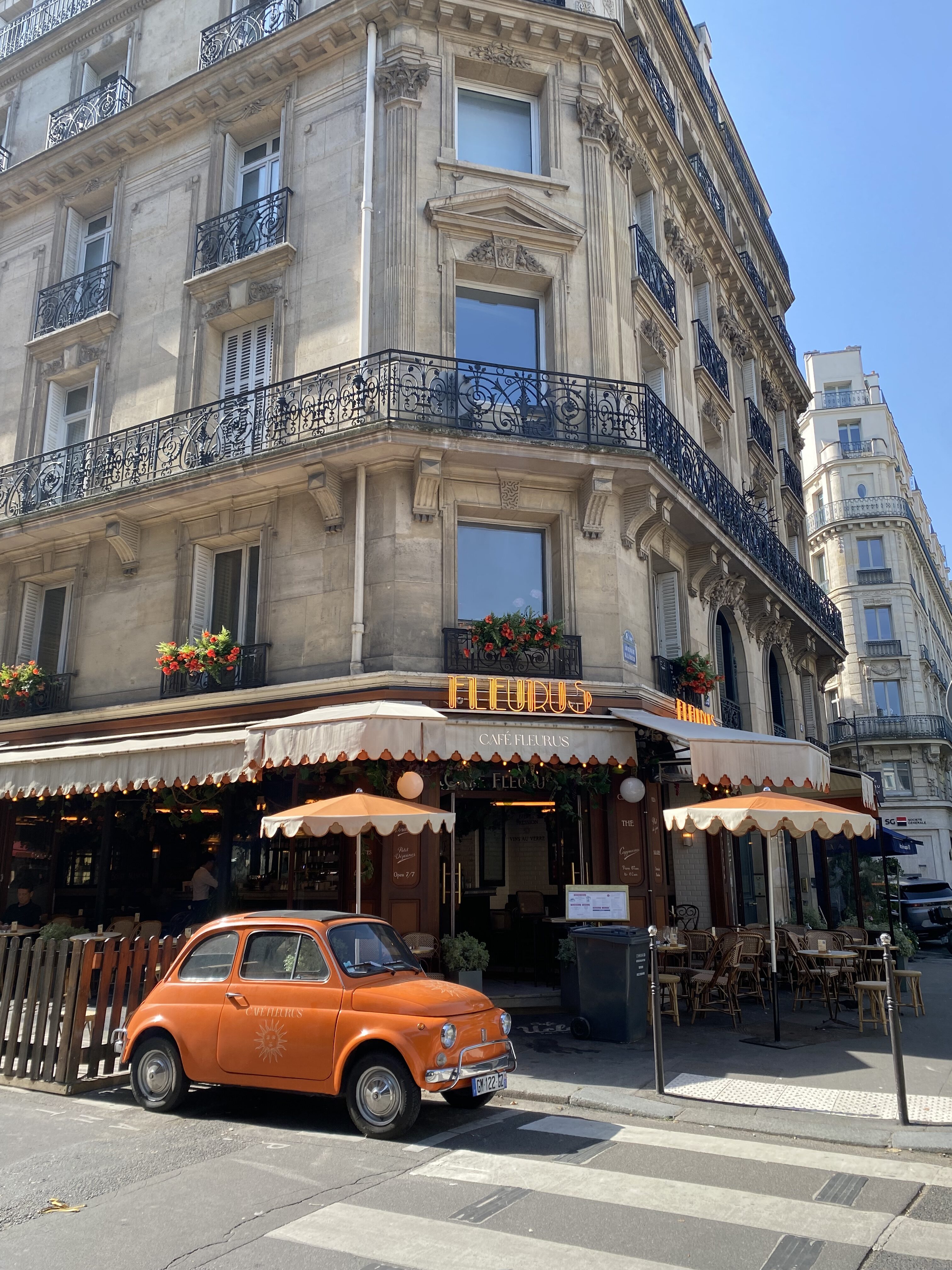 A charming Parisian cafe showcasing traditional Haussmann architecture with orange umbrellas and a vintage, bright orange car parked on the adjacent street.