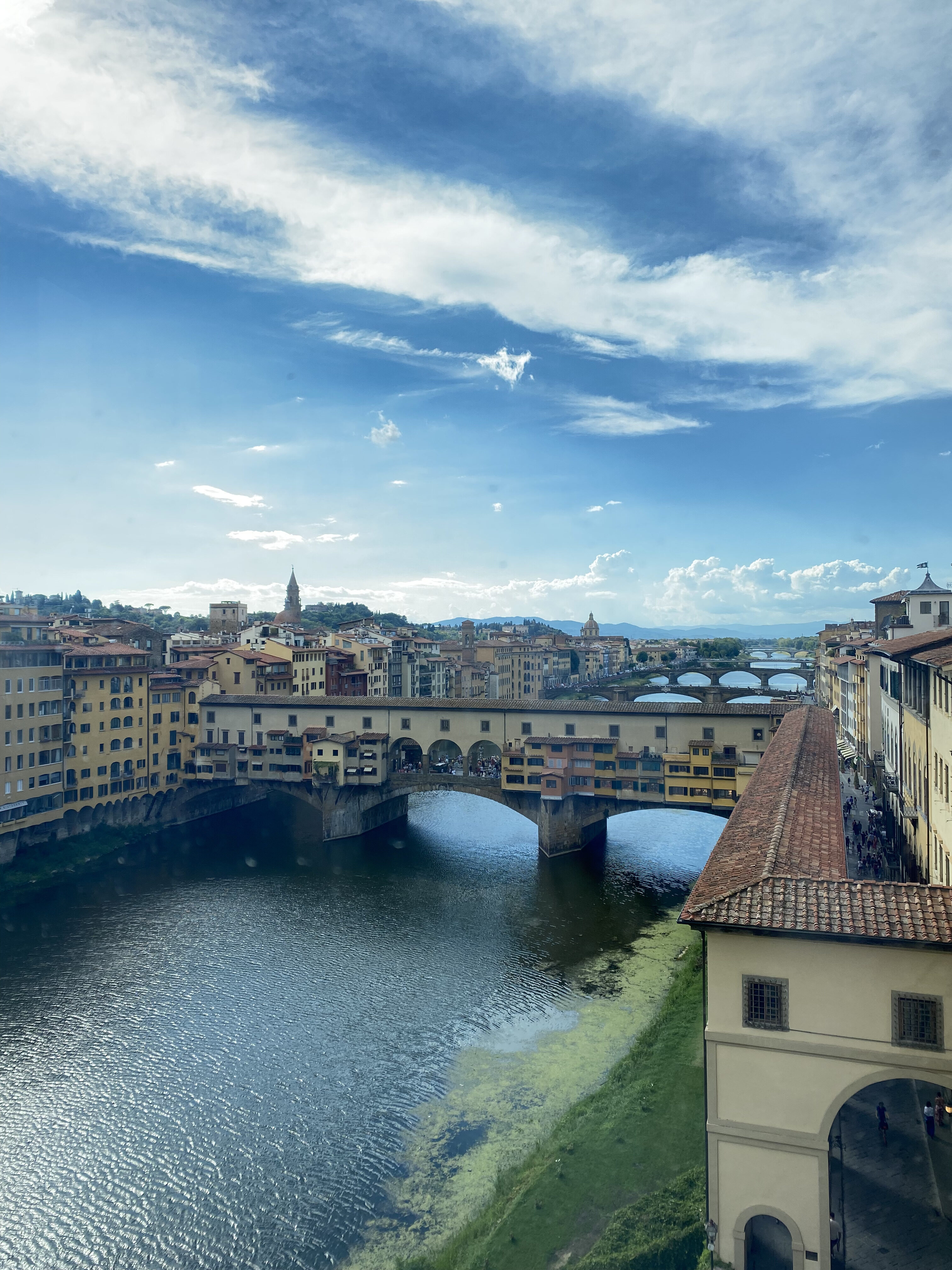 The Ponte Vecchio and the Arno River seen from a window at the Uffizi Gallery. A bright, cloudy blue sky paints the horizon.