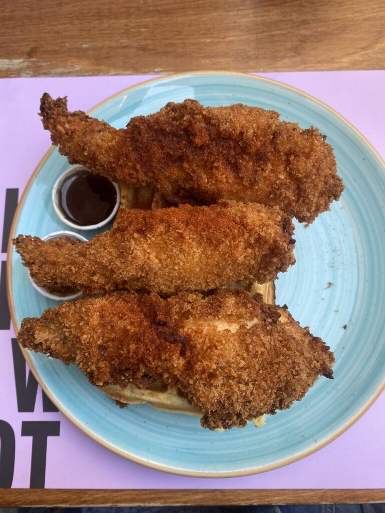 A perfectly-fried plate of chicken in waffles with three chicken tenders stacked atop a waffle on a blue plate. A pink table map sits beneath the plate onto of a wooden table.