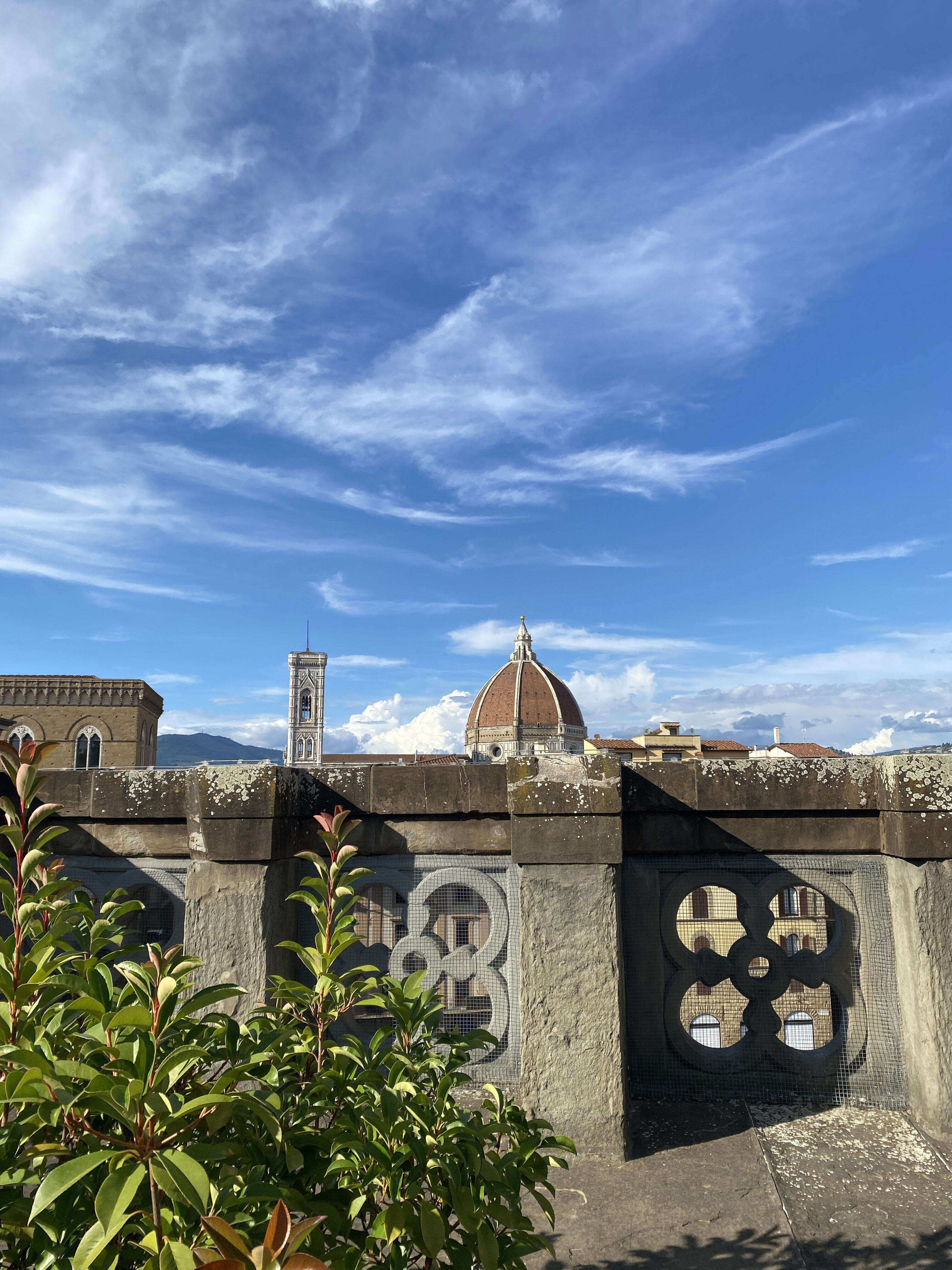 Brunelleschi's duomo peeks over the concrete decorate railing at a rooftop cafe at the Uffizi Gallery. A bright blue sky paints the horizon.