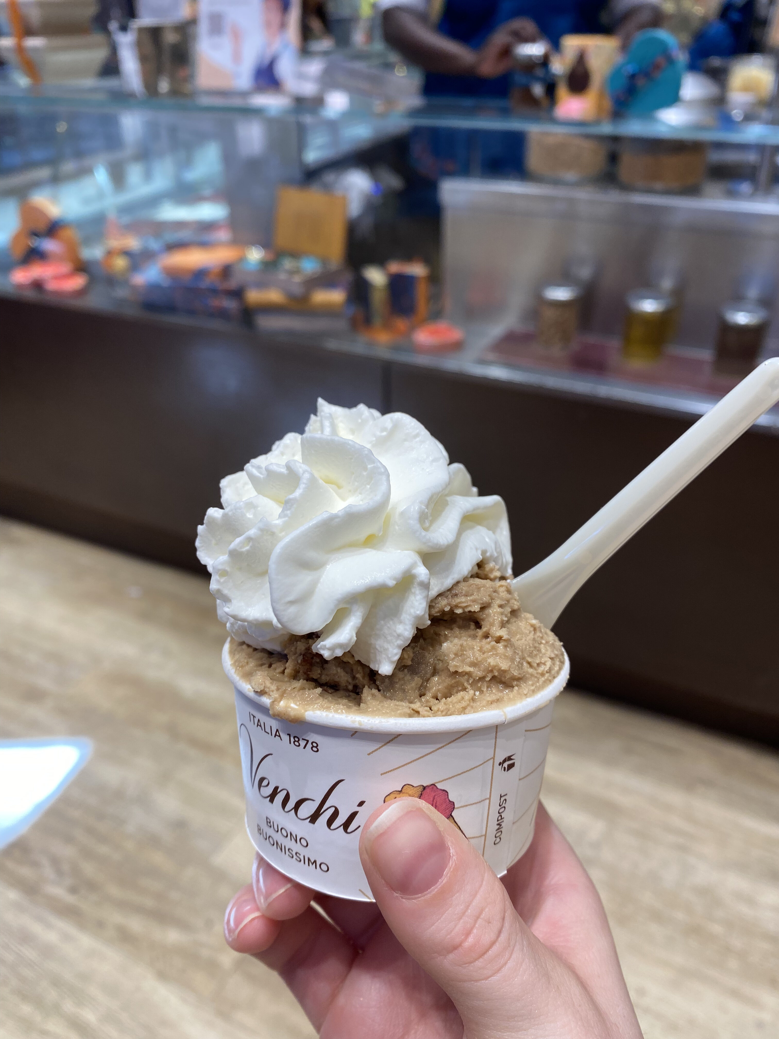 A caramel-colored gelato in a cup, topped with fluffy whipped cream with a spoon. In the background is a display of gelato flavors. The gelato cup reads, "Venchi".