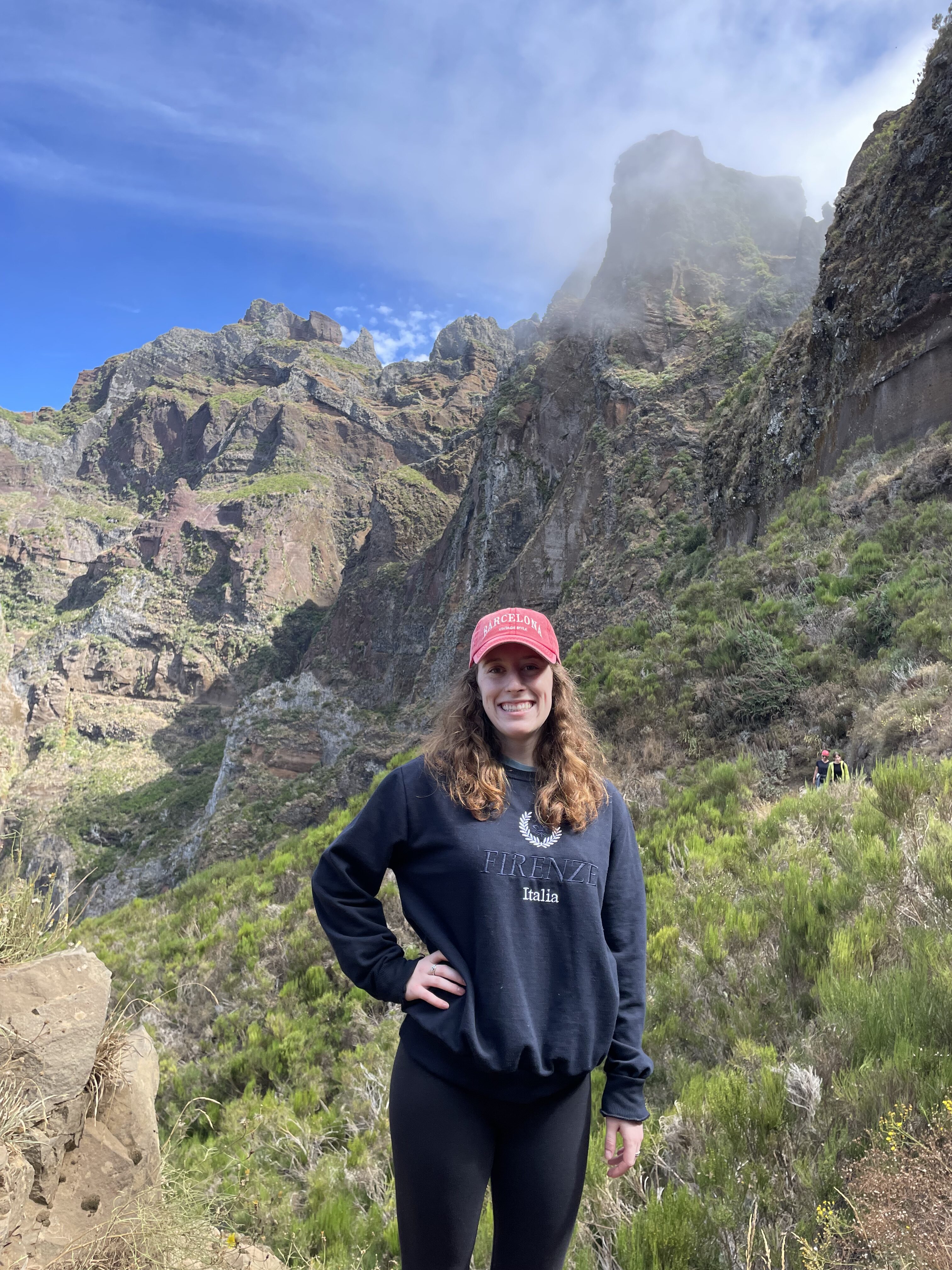 Bella, wearing a blue sweatshirt and a pink hat, poses in front of a jagged mountain along the Pico Ruivo hike in Madeira.