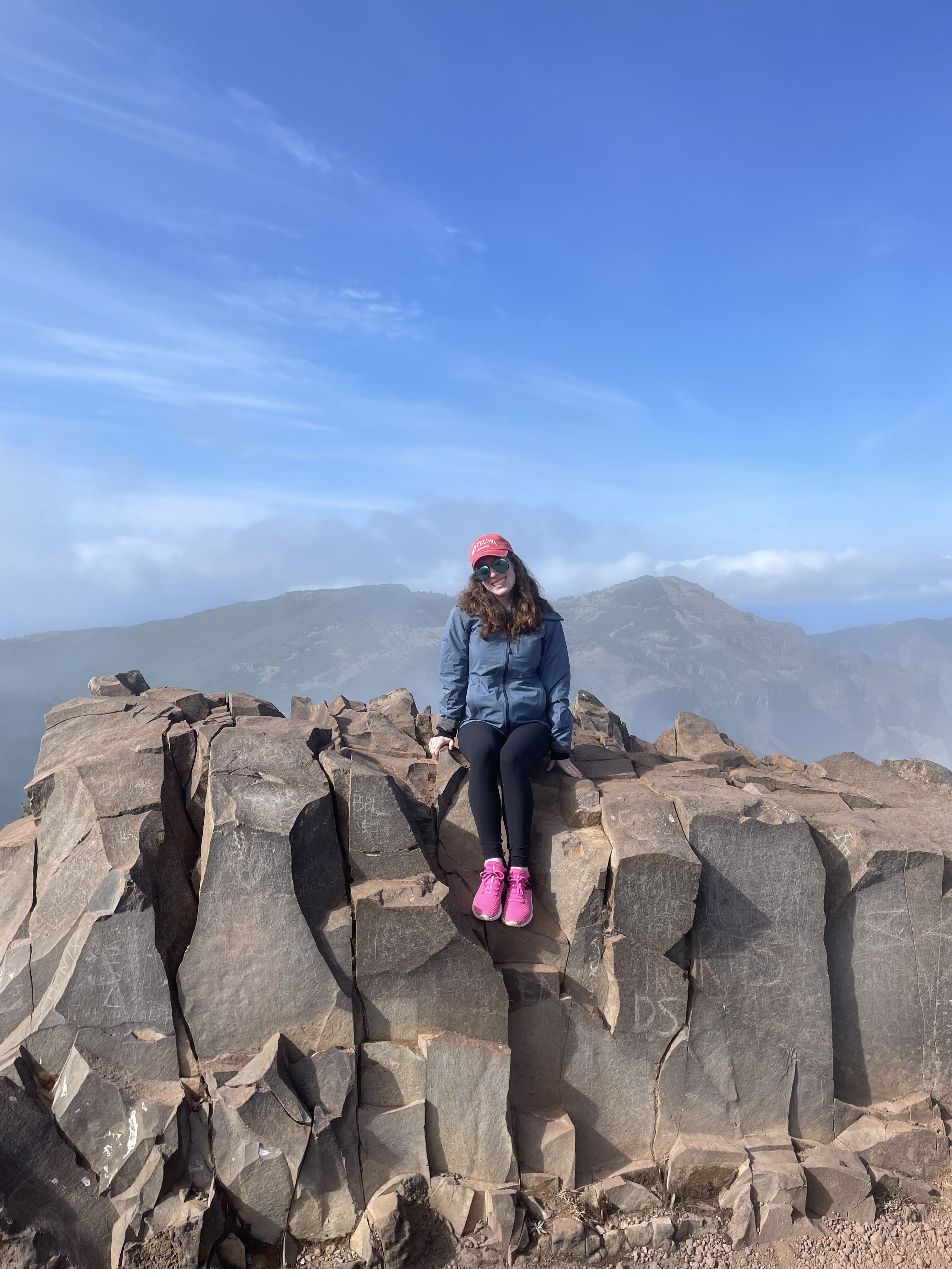 Bella sitting on a jagged rock formation along the Pico Ruivo hike in Madeira. A bright blue sky frames the background.