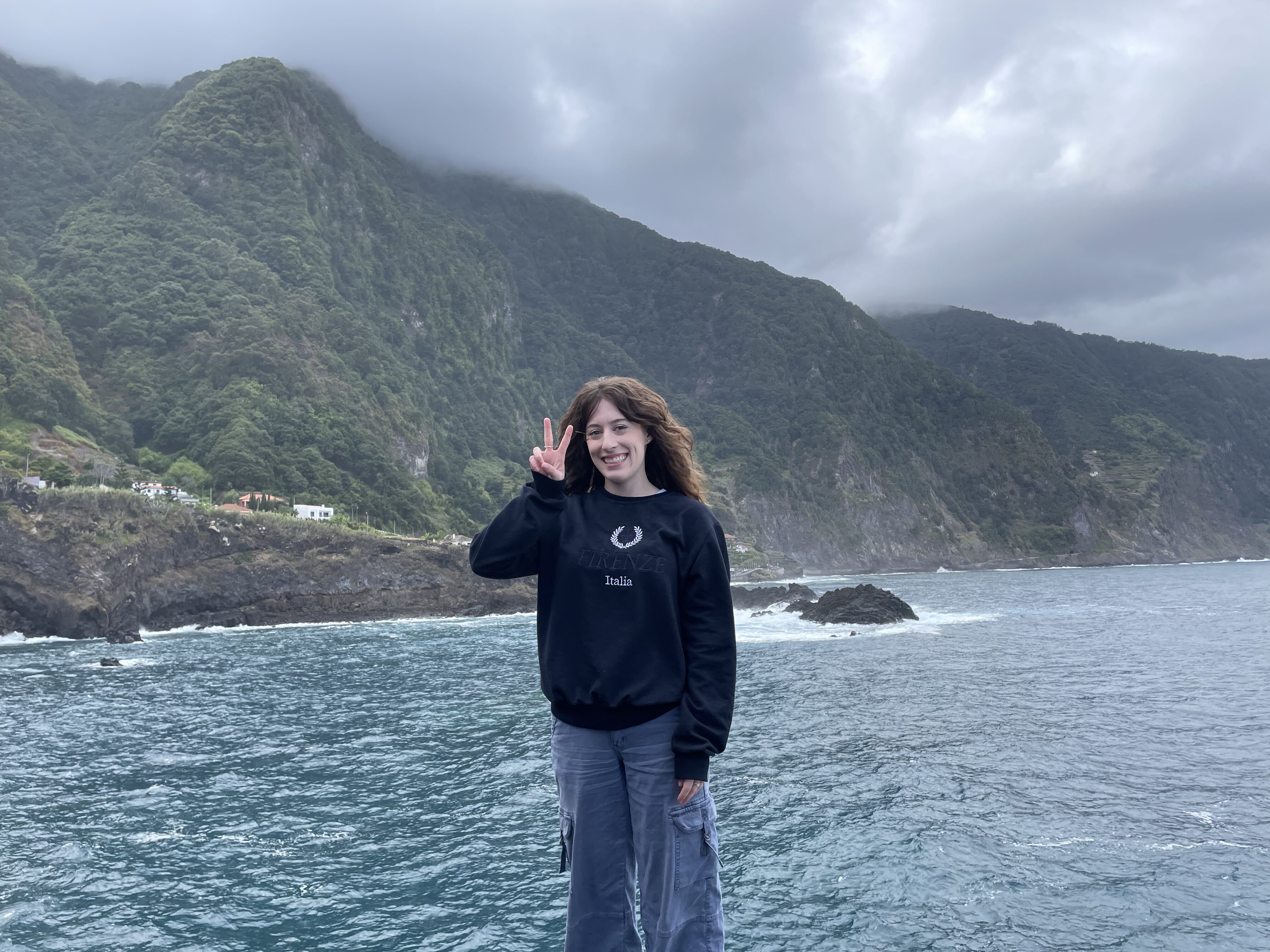 Bella holding up a peace sign in front a the blue ocean and the green mountains of Madeira.