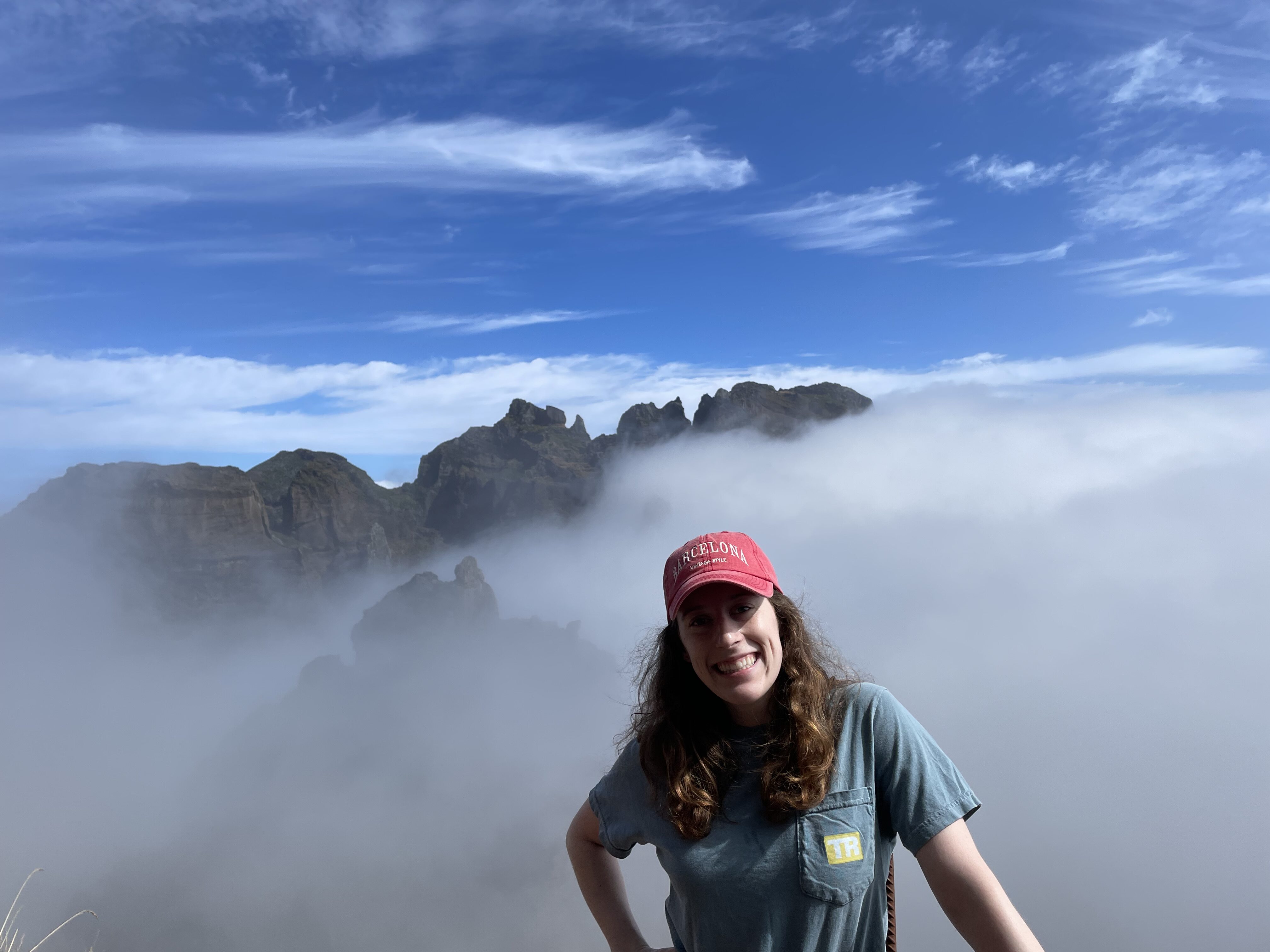 Bella, wearing a bright pink hat and a blue shirt, smiles in front of a cloud-covered mountaintop in Madeira.