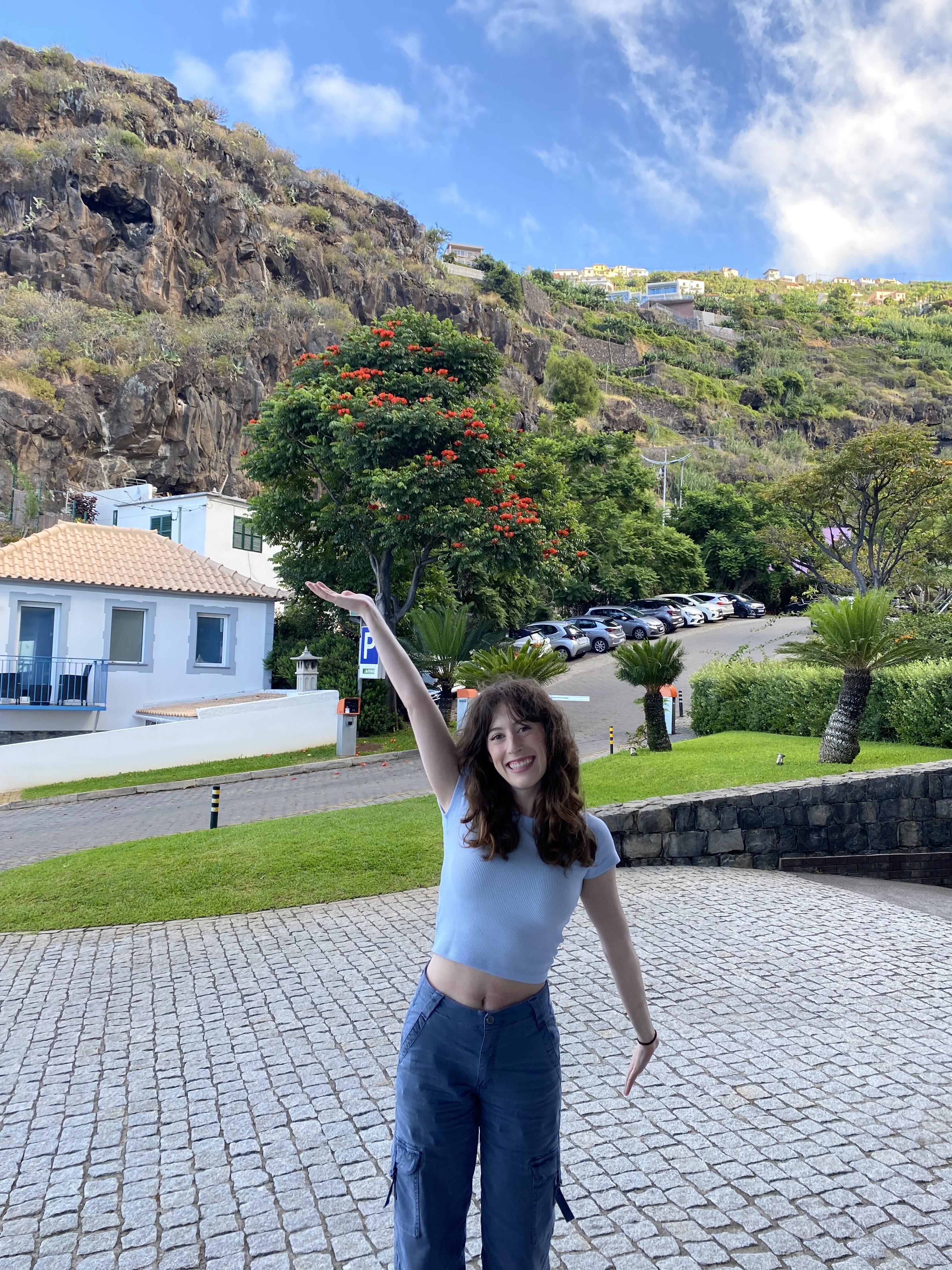 Bella, poses with one arm up and one arm down, in front of a hillside in Madeira.