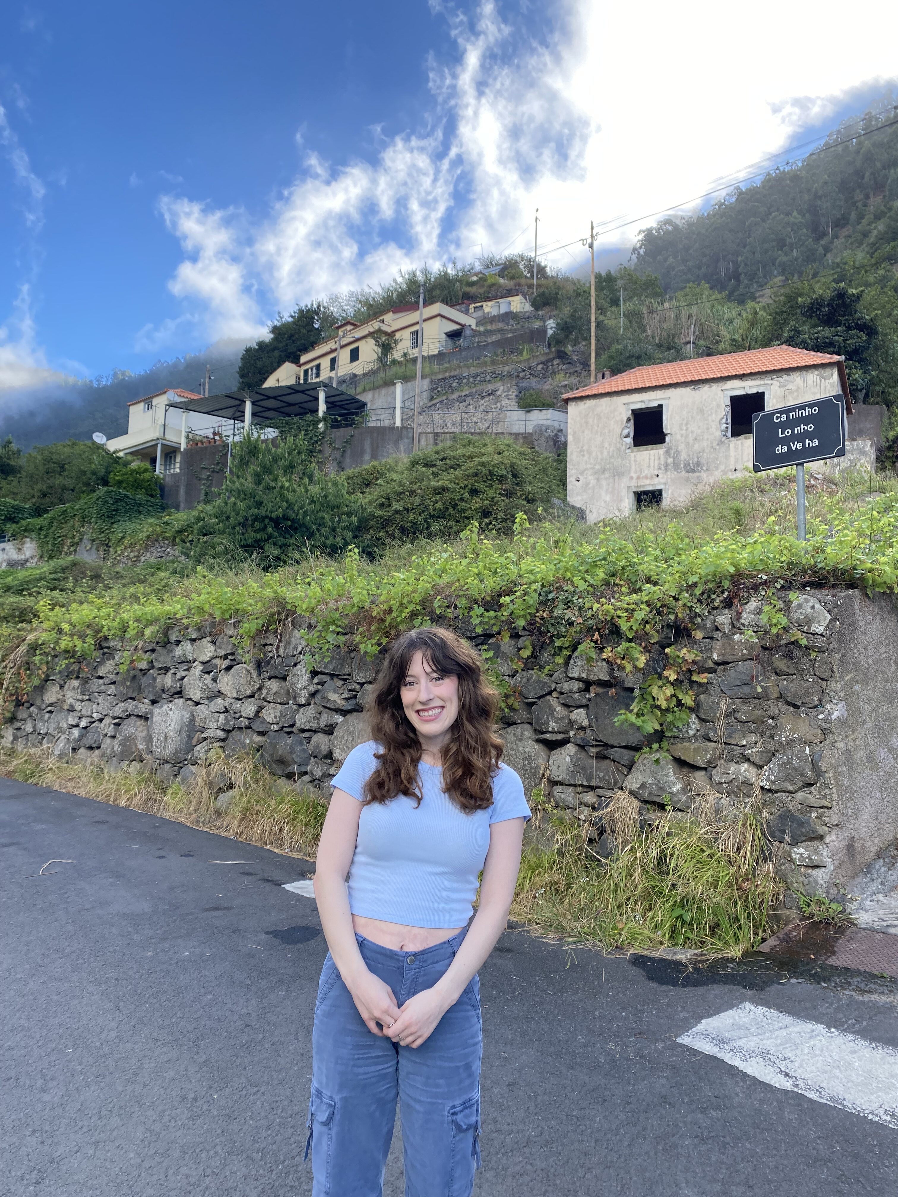 Bella, wearing a light blue crop top and blue cargo pants smiles in front of a hillside village in the mountains of Madeira.