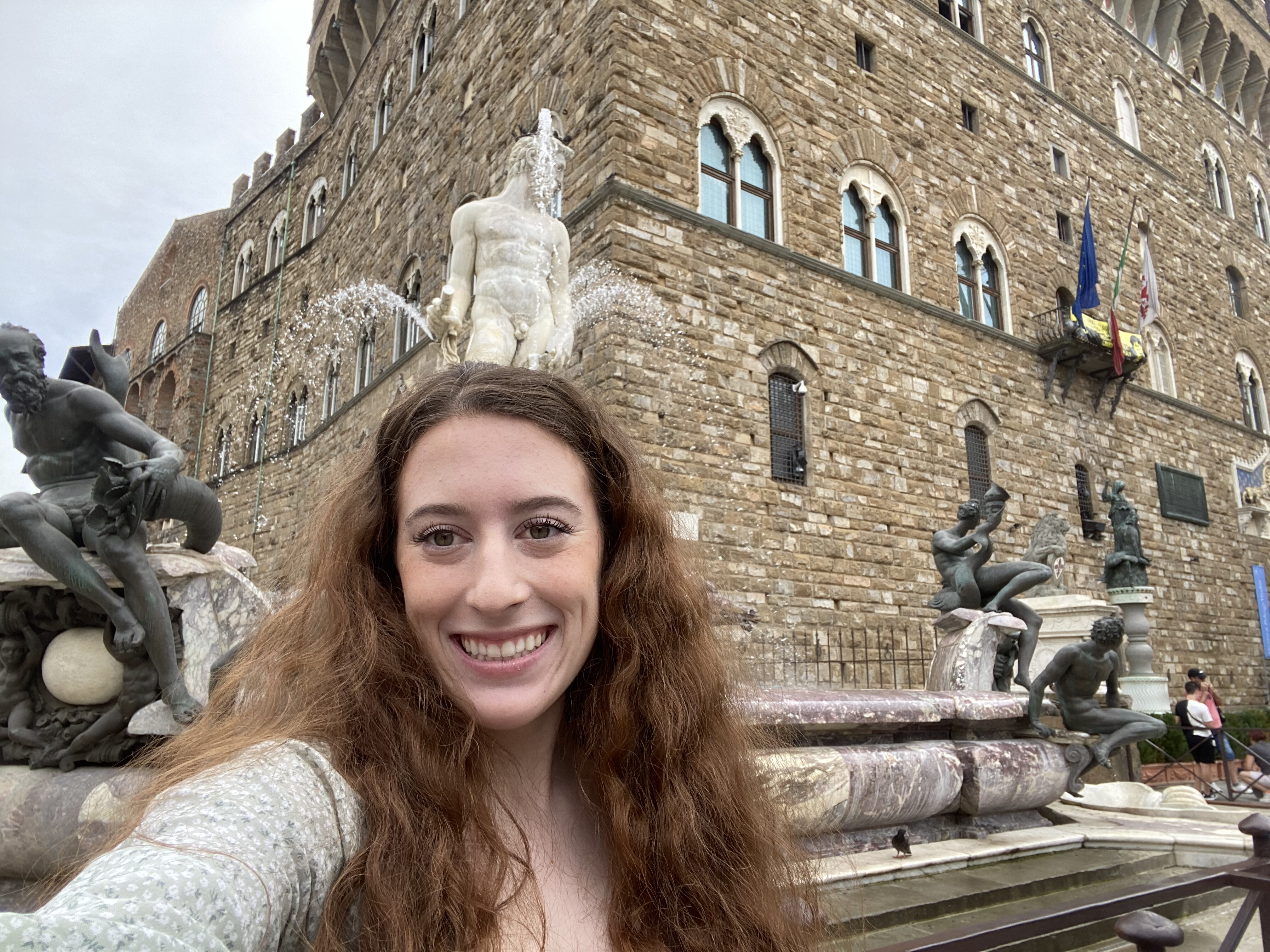 Bella standing in front of a white marble statue towering over a fountain, flanked by several bronze statues. A weathered stone building, Palazzo Vecchio, stands in the background.