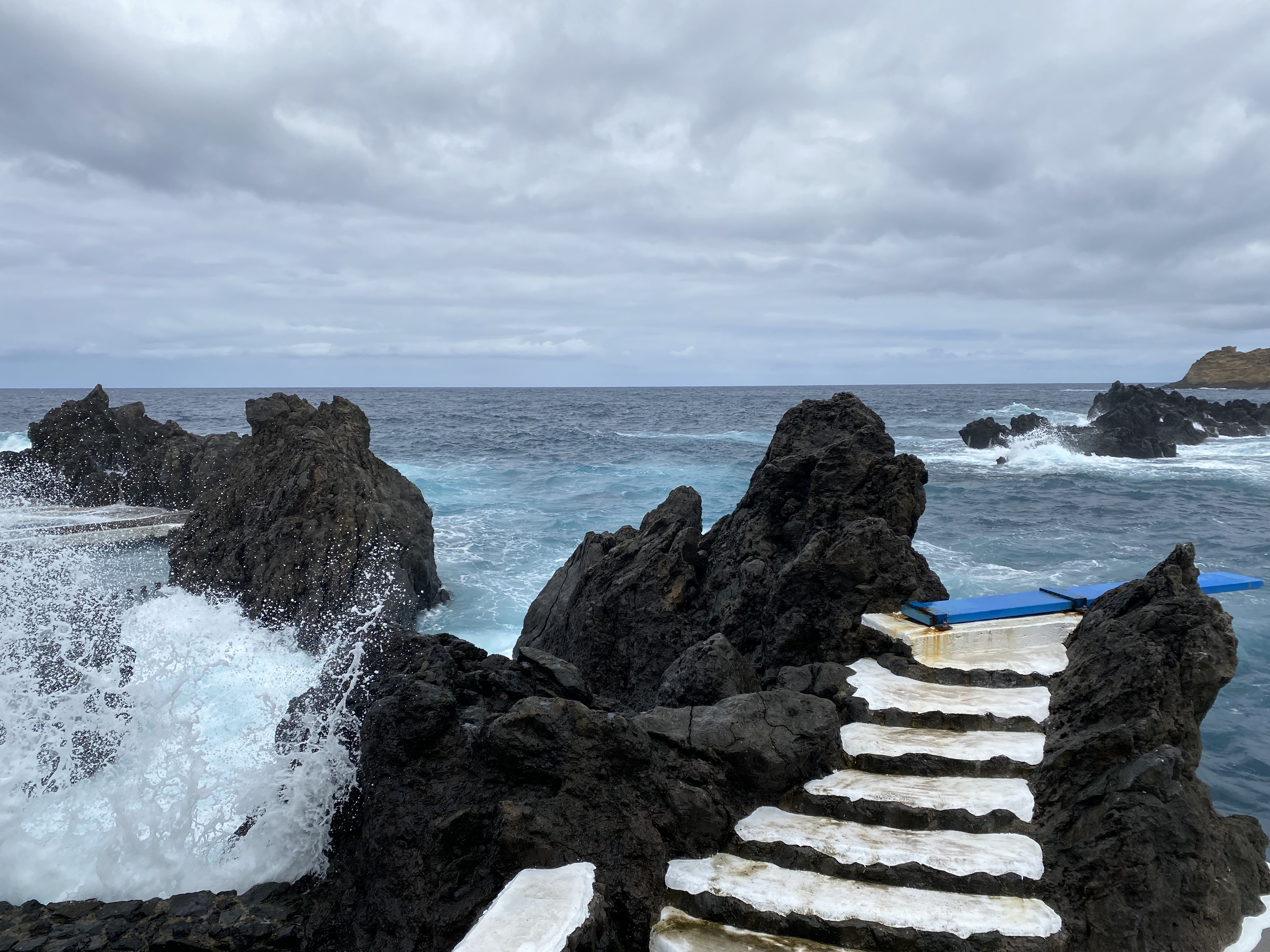 Blue diving board with white steps between a rock formation in the Porto Moniz pools