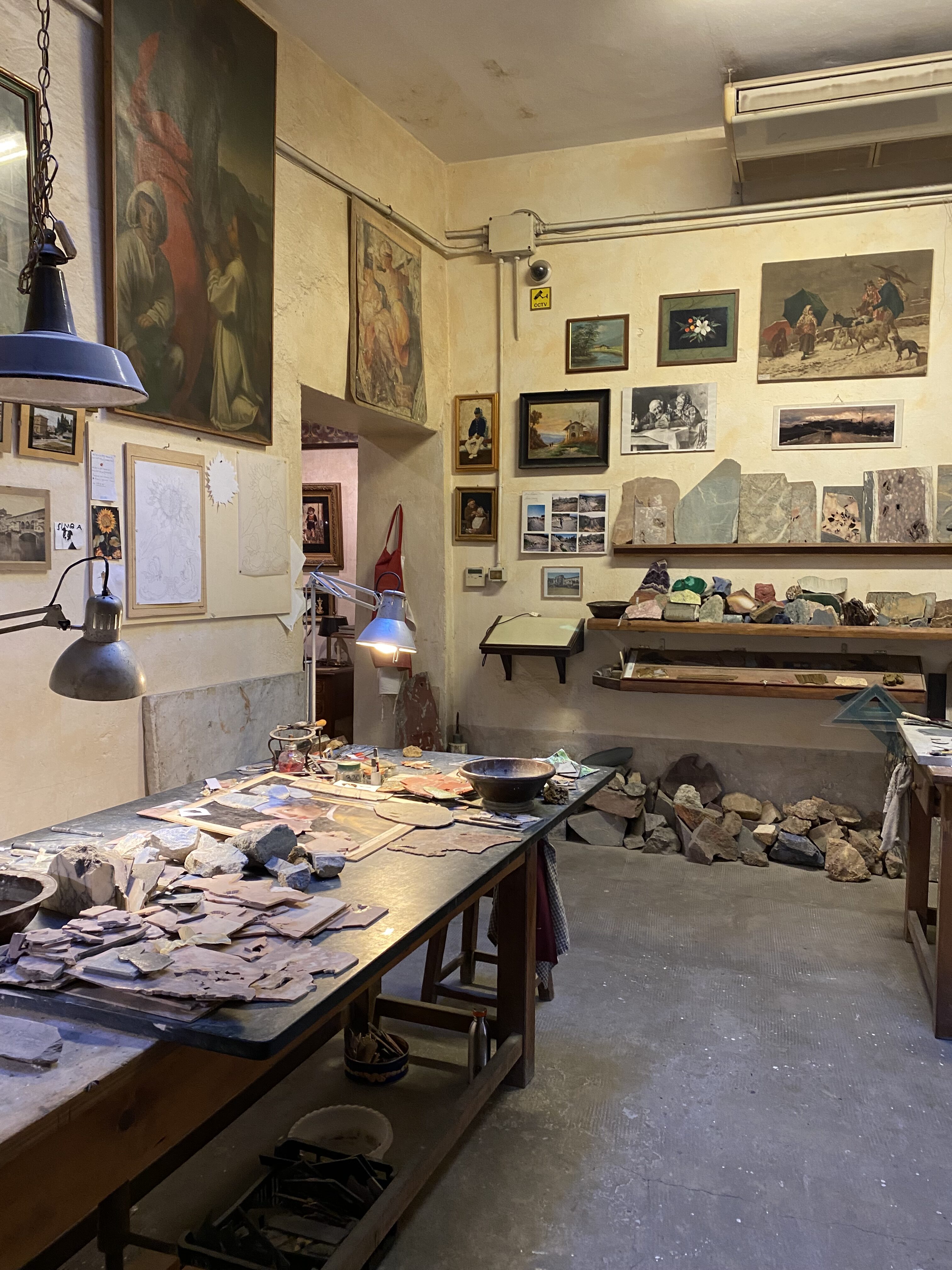 A local mosaic workshop in Florence with a table decorate with uncut stones of all colors. Posters on the wall show replicas of famous artwork.