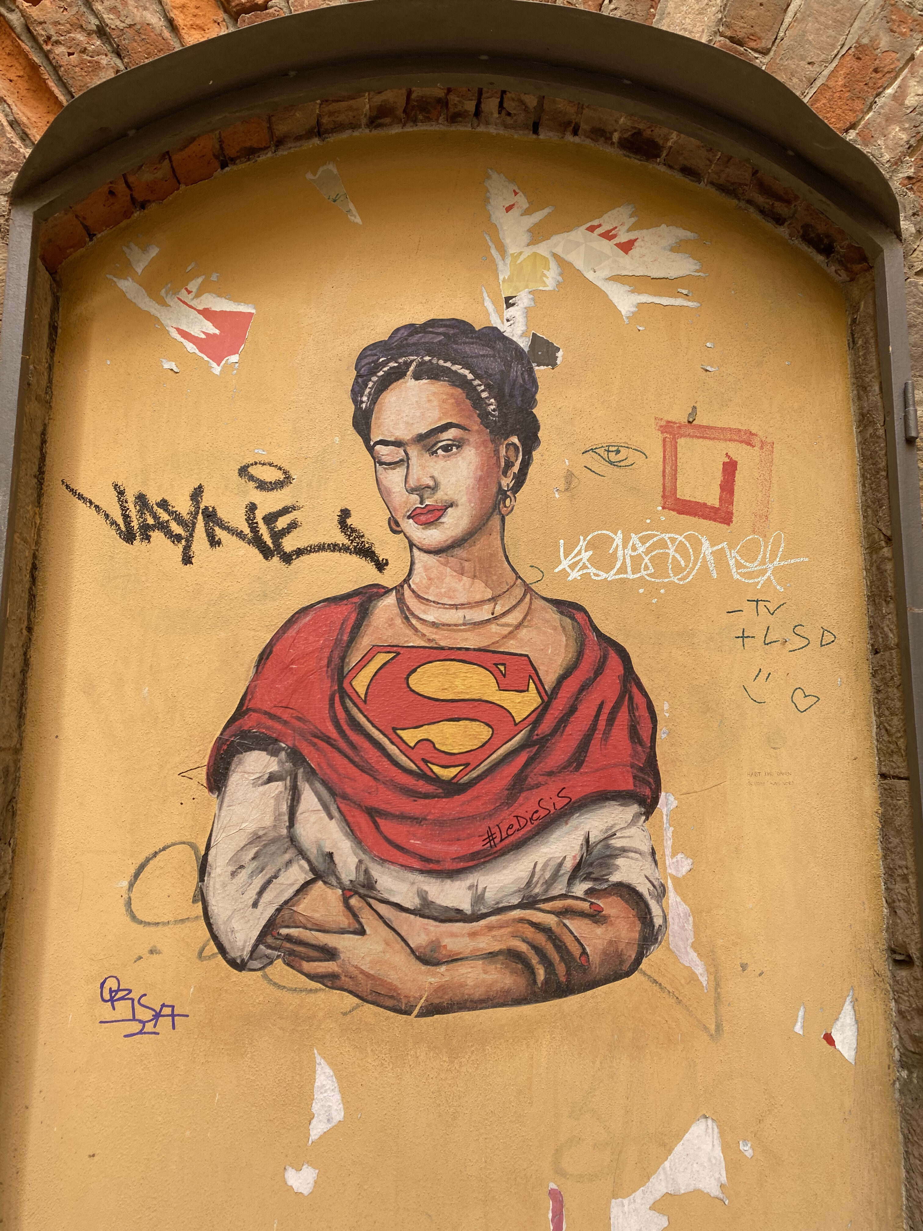 A street art portrait on a yellow wall of Frida Kahlo wearing red shawl that unravels to reveal the Superman logo across her chest.