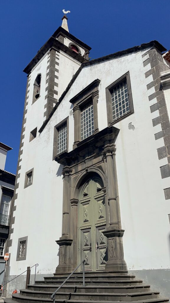 A white and dark stone building with a monumental dark stone door in front of a dark blue sky in Funchal Old town.