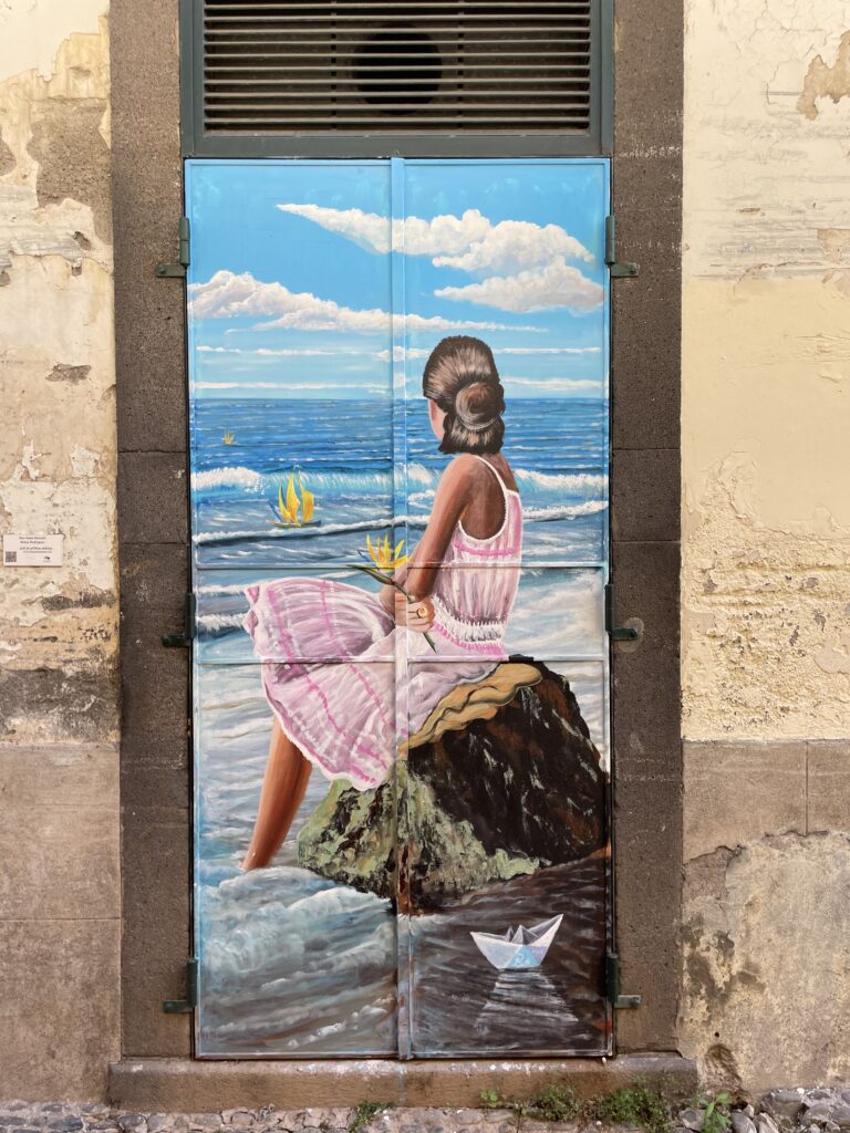 Funchal street art showing a young tan women in a pink dress starting out to sea while sitting on a rock.