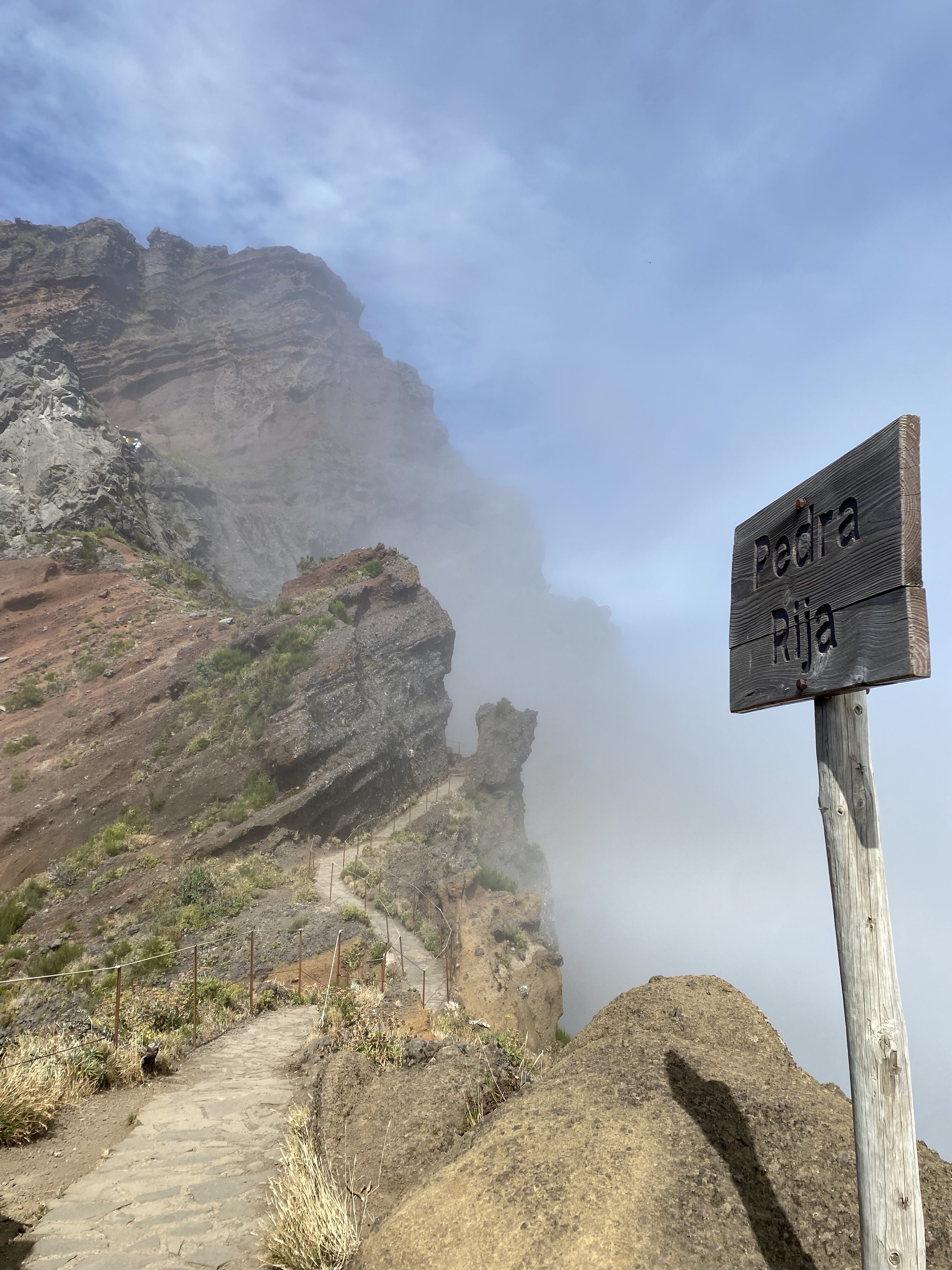 A Rocky Mountain trail in Madeira with a rickety wooden sign pointing towards the ascent. The sign reads, "Pedra Rita", which is a viewpoint overlooking the mountains in Madeira.