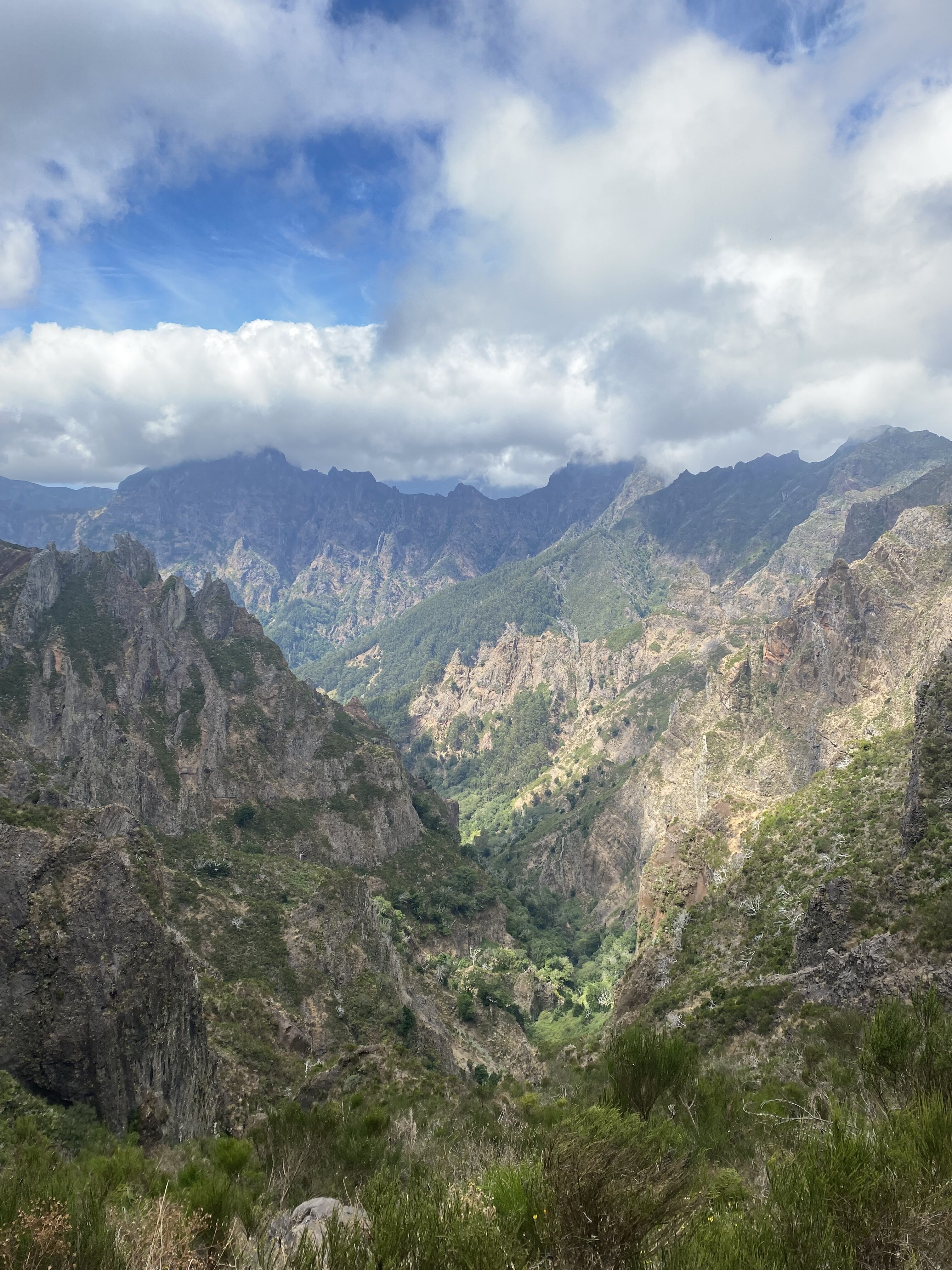 A beautiful range of mountains in Madeira with tan rocky faces and vibrant green vegetation. A cloudy blue sky casts shadows over some of the peaks.