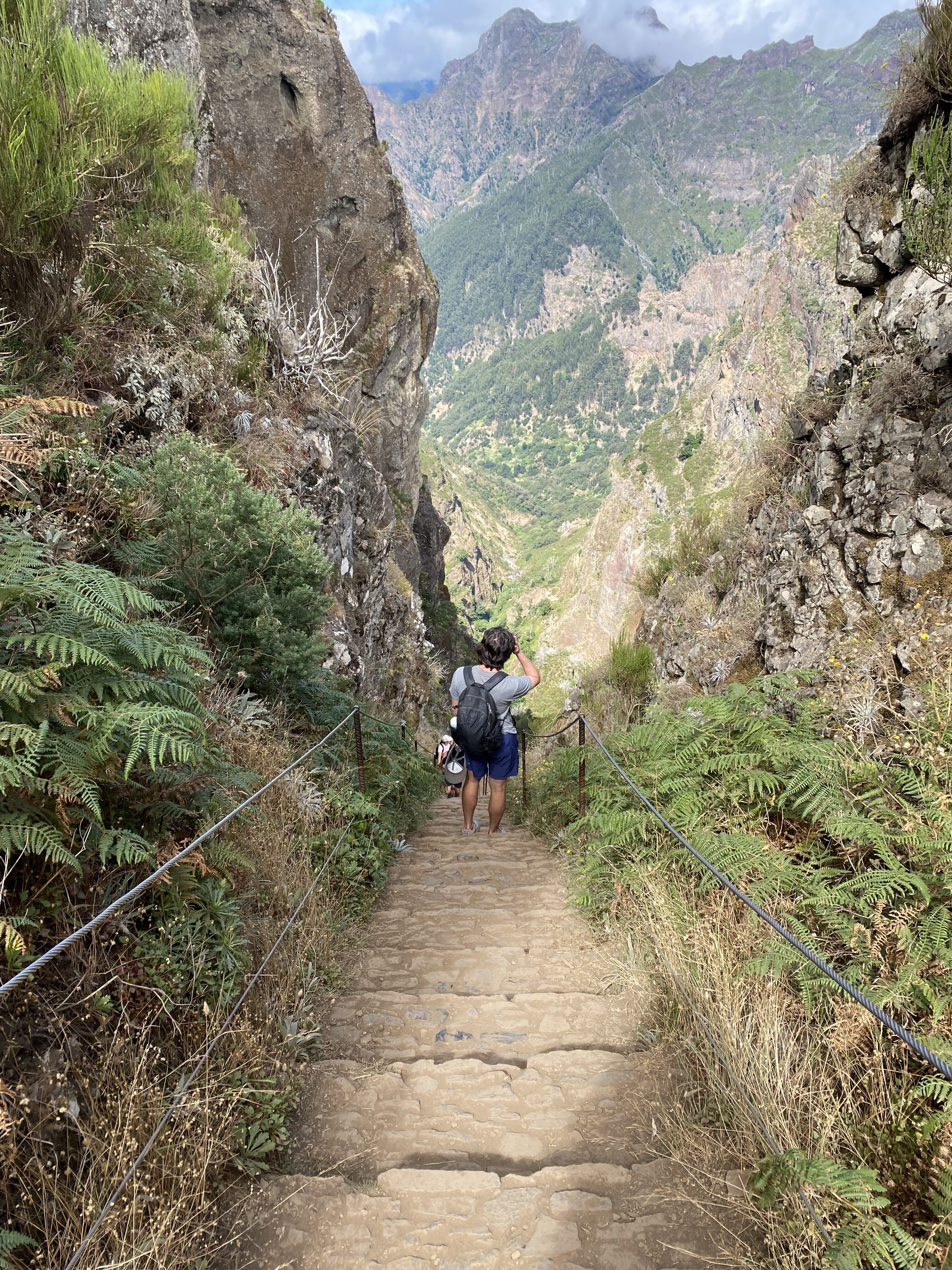 A steep set of stairs along the Pico Ruivo hike with rock formations on either side. At the bottom of the stairs, a man wearing a backpack admires the view.