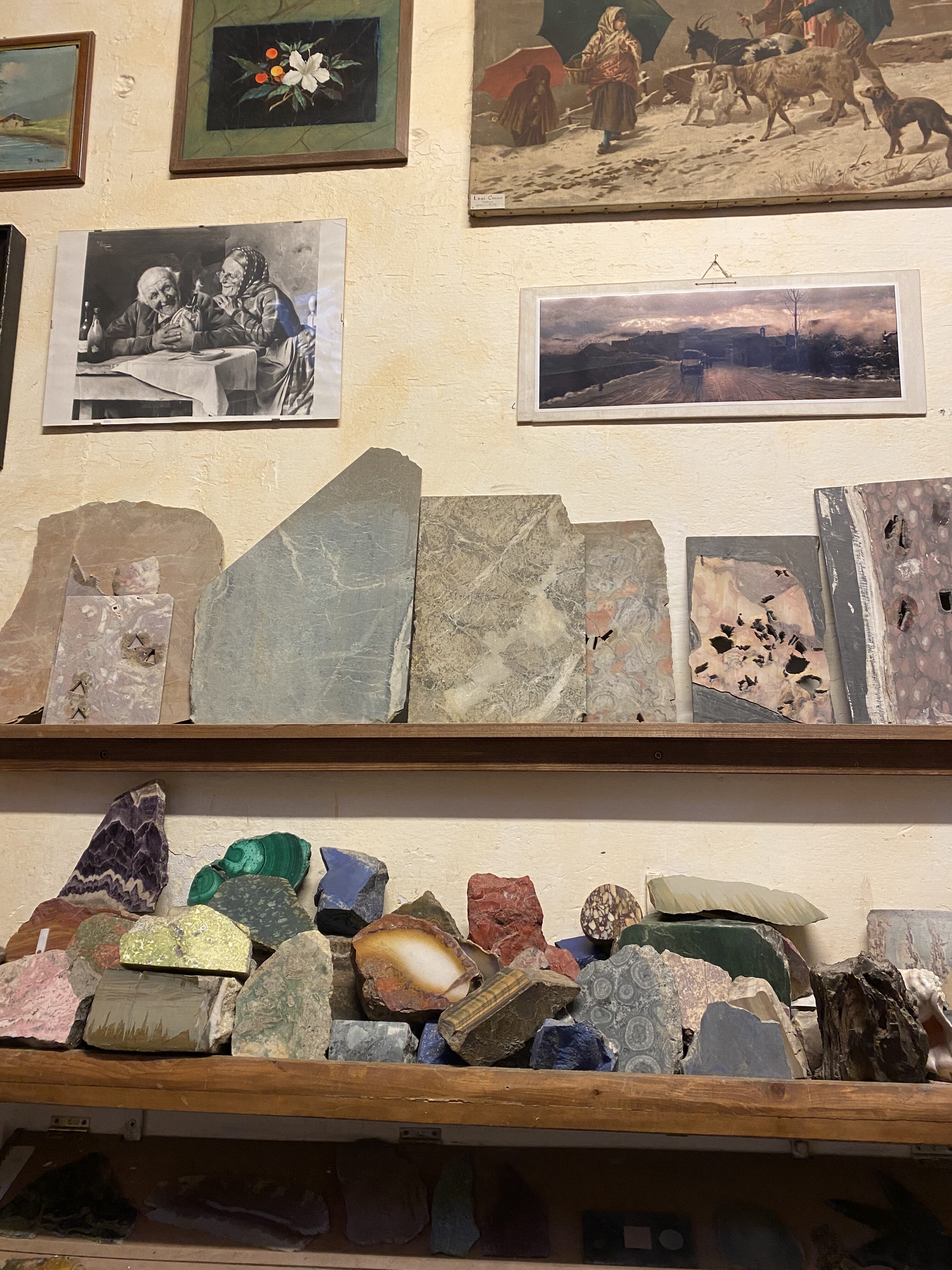 Unpolished stones placed on a shelf along a tan wall with various small posters containing artwork surrounding the stones.
