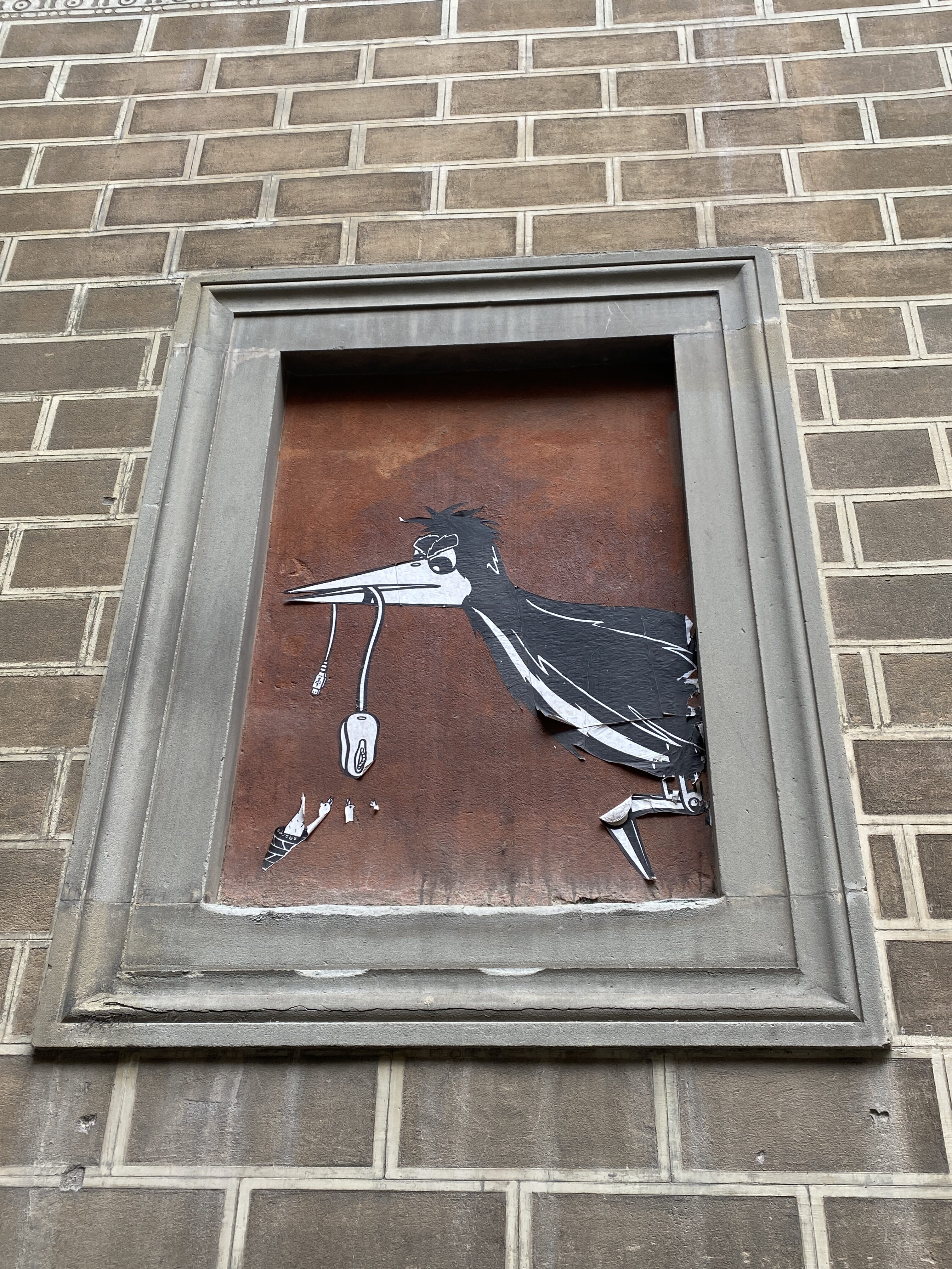 Framed street art set against a brown, weather brick wall showcasing a black bird dangling a computer mouse over its nest of baby birds.